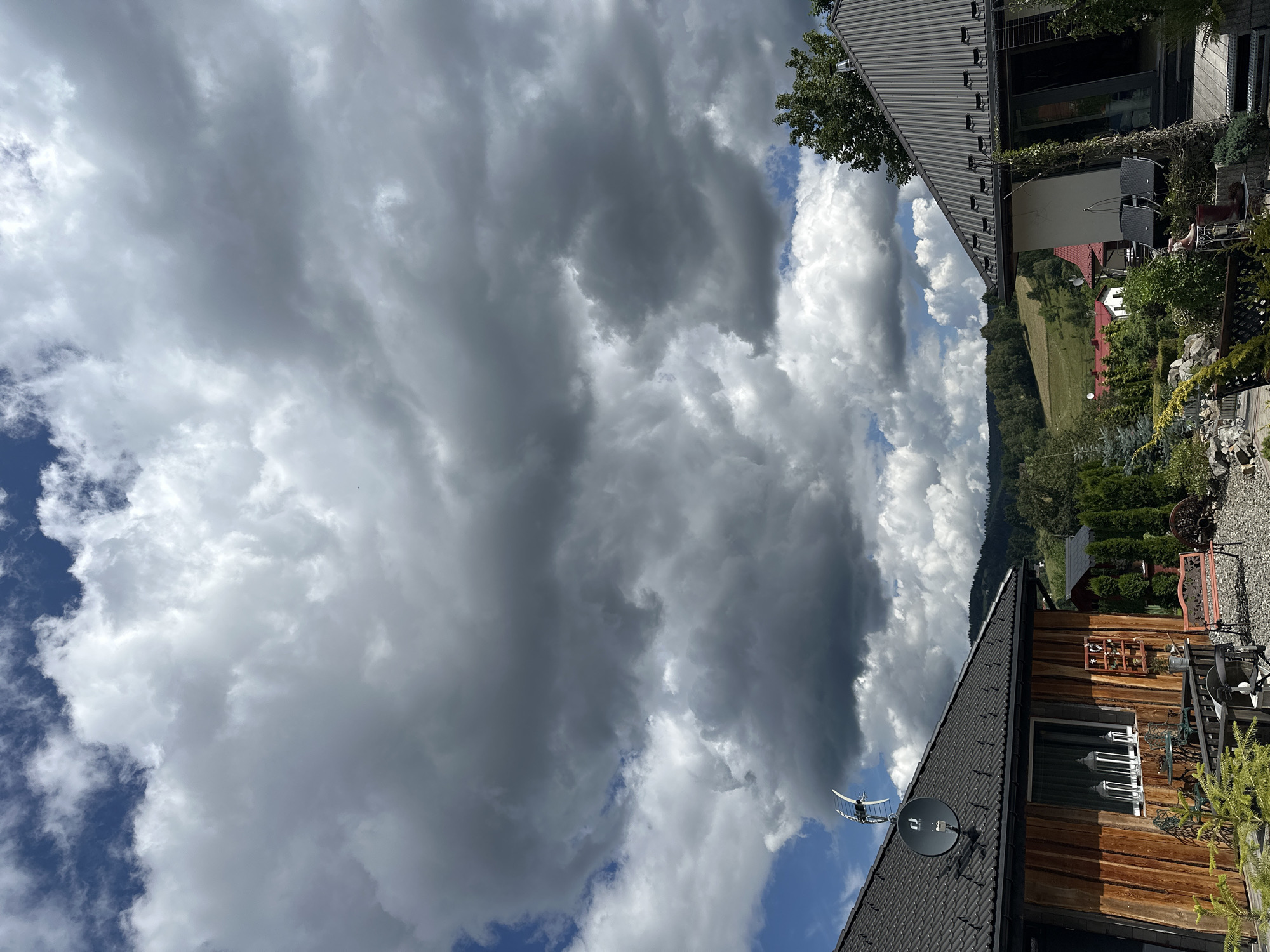 The image depicts a partly cloudy sky over a suburban area in Wisła, Poland. Two houses with dark-colored roofs are visible in the foreground. The house on the left has a wooden facade and a satellite dish mounted on its roof. The house on the right features a white facade and a modern, dark metal roof. In the background, rolling hills covered with trees and some red-roofed buildings can be seen. The environment appears to be a residential area with a well-maintained yard in front of the houses, featuring various plants, outdoor furniture, and gravel pathways. There are no people visible in the scene.