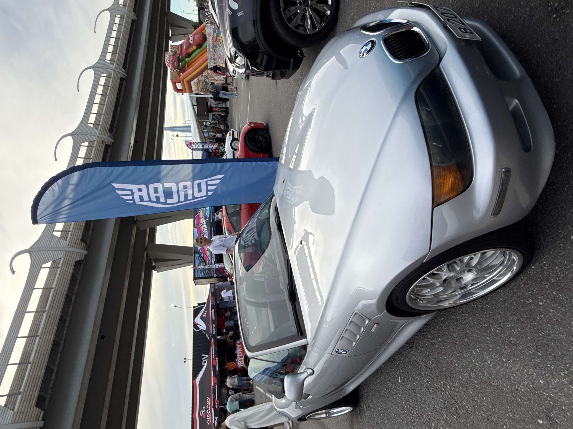 A silver BMW Z3 convertible is parked on asphalt, facing slightly left, under a large overpass. The car has a black interior and a black license plate with white lettering that reads "B 207". In the background, several tents are set up, some displaying logos. A blue banner with white text that appears to be "RADCAGE" stands to the right of the car. Behind the tents, an inflatable slide with cartoon characters is visible. A black SUV is parked to the right of the BMW. People are gathered around the tents and near the vehicles. The scene is likely taking place at an outdoor event or car show, possibly in St. Petersburg, Russia, given the visible architecture of the overpass. The sky is overcast.