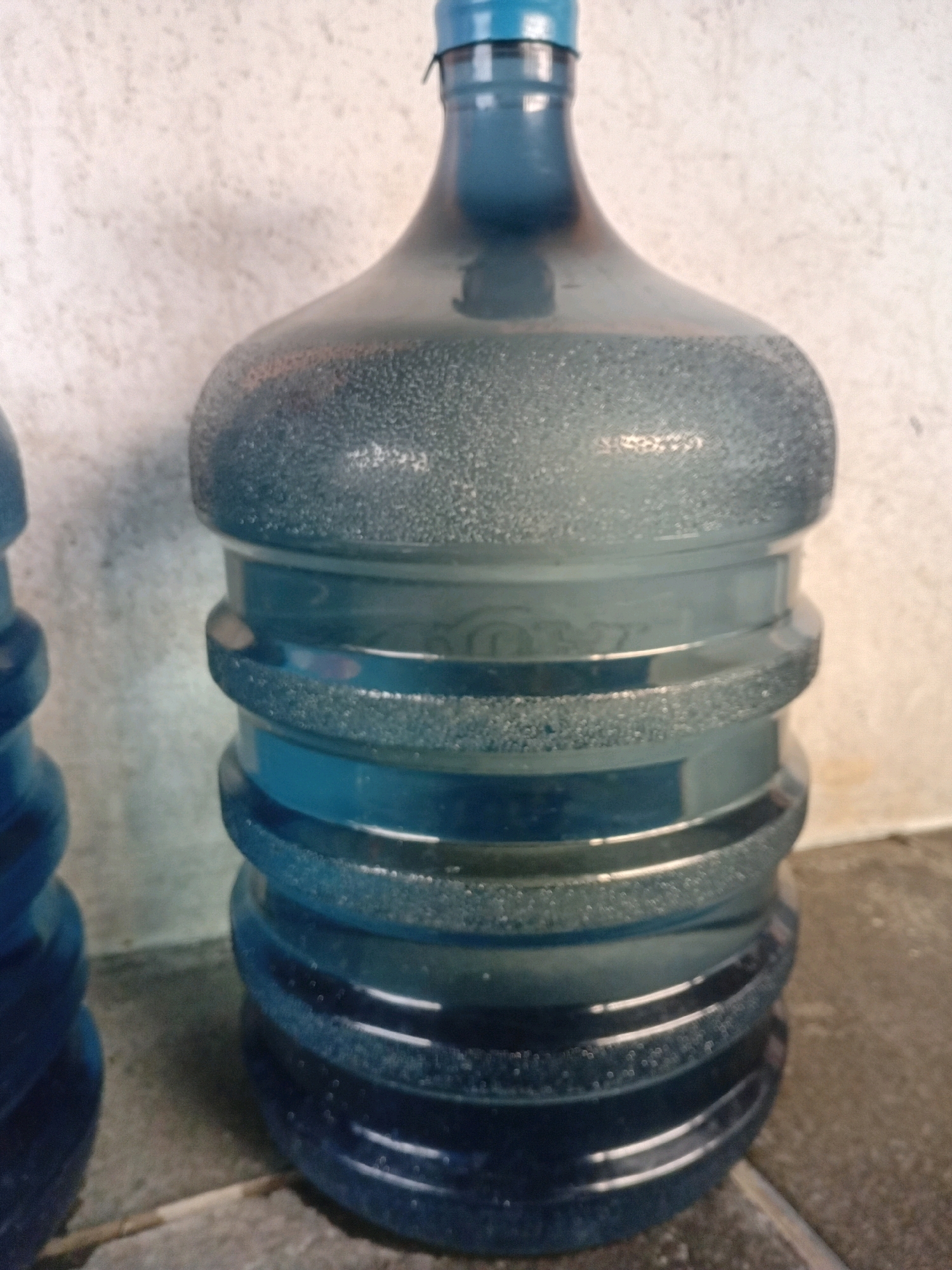 A blue plastic water jug is shown in close-up, filled with water and resting on a tiled floor. The jug has a narrow neck topped with a blue cap, and its body is segmented by raised, textured bands. Tiny bubbles are visible within the water, clinging to the inner surface of the jug and dispersed throughout the liquid. The background is a textured, light-colored wall. Another blue plastic object, also appearing to be a water jug, is partially visible to the left of the main subject. The lighting suggests an indoor environment. The scene is likely from a residential or commercial area within Tangerang, Indonesia, a city known for its urban landscape and access to bottled water supplies.