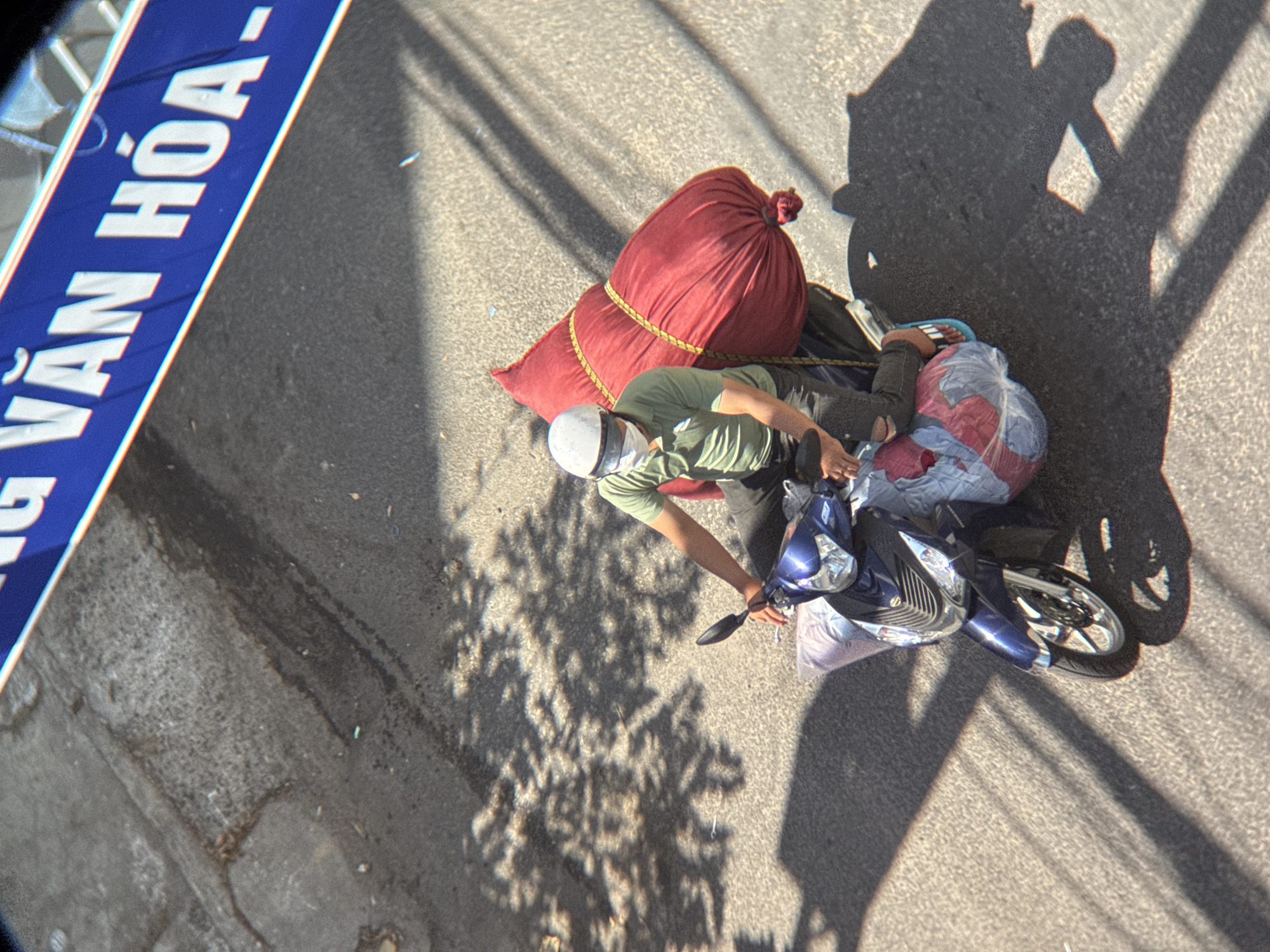 In Nha Trang, Vietnam, a person wearing a white helmet, a face mask, and a green t-shirt rides a blue motorcycle. The motorcycle is loaded with several large, red and white fabric bags. The person is seated on the motorcycle, with their legs extended and their feet wearing sandals. The motorcycle is on a paved road, with shadows of trees cast across the surface. A blue sign with white text is visible in the upper part of the image.