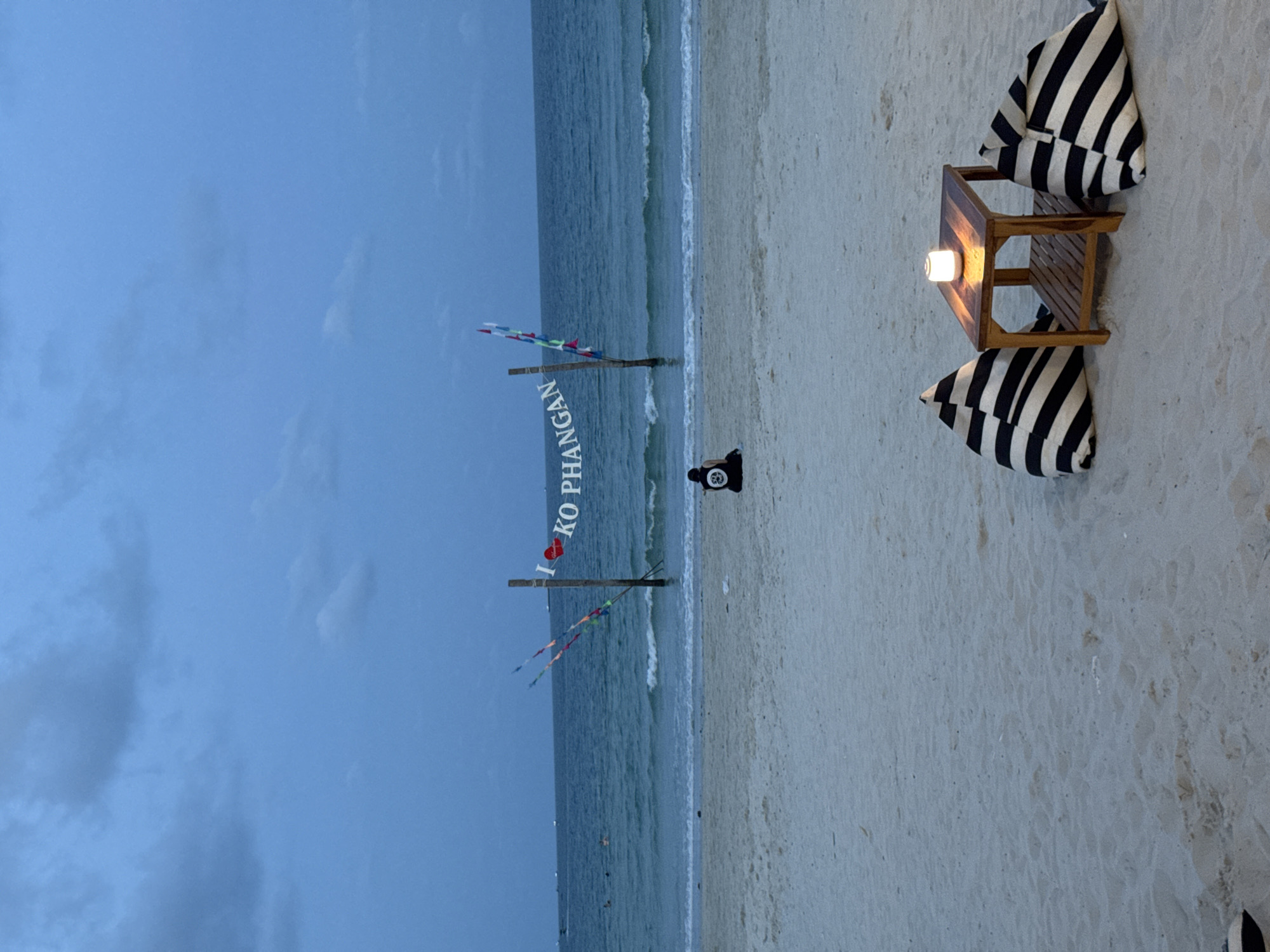 Here is a description of the image.
The image depicts a beach scene, most likely in the evening, as indicated by the dim lighting and the presence of a lamp lit on a small table. The beach has white sand, and the sea appears calm with gentle waves. A sign reading "I <3 KO PHANGAN" is visible on wooden posts adorned with small colorful flags. A person is visible near the sign, seemingly standing or sitting on the beach. To the right, there is a small wooden table illuminated by a lamp. Two striped cushions are arranged around the table, suggesting a seating arrangement for two. The sky is cloudy, with a deep blue hue. The image gives the impression of a tranquil and romantic setting on the beach. It is probably located in Ban Haad Rin, Thailand, based on the text on the sign "KO PHANGAN".