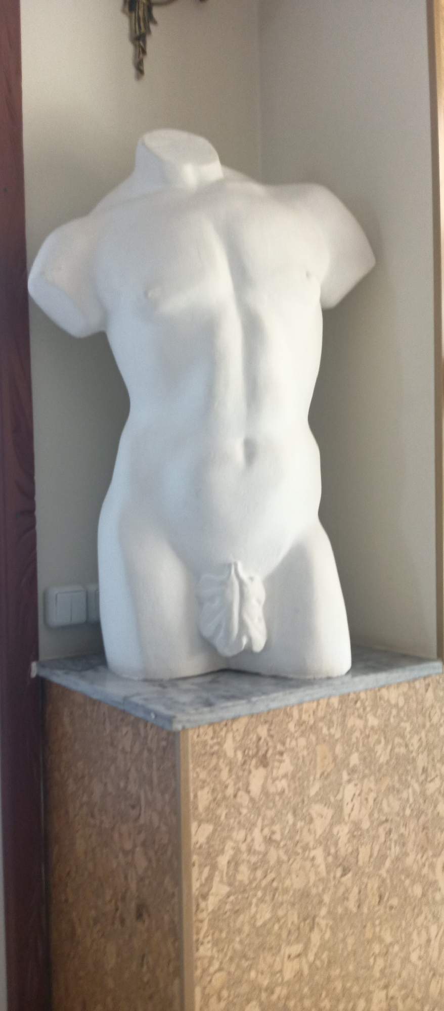 The image shows a white male torso statue, lacking arms, legs, and a head, prominently displayed on a gray marble surface atop a wooden pedestal. The statue has an abdomen, a navel, and a penis with leaves. The backdrop consists of off-white walls, with what appears to be an electrical outlet visible to the left of the statue's base. A partially visible light fixture is at the upper part of the image, and a doorframe is on the left edge. The environment suggests the statue is in a niche or display area within a building. The location is Unknown, Unknown.