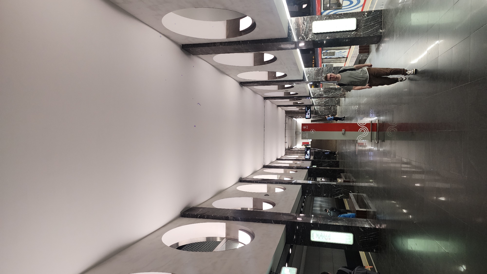 The image depicts a modern interior space, likely a subway station in Moscow, Russia, characterized by clean lines and a minimalist aesthetic. The station is adorned with gray marble and white walls with circular openings. A man stands on the polished black tile floor, facing the camera, wearing a gray t-shirt, brown pants, and sneakers, with a backpack slung over his shoulder. Other indistinct figures are present in the background. Illuminated signs are visible. A subway car can be seen beyond the man. A red informational panel featuring Moscow Metro branding is visible in the midground.