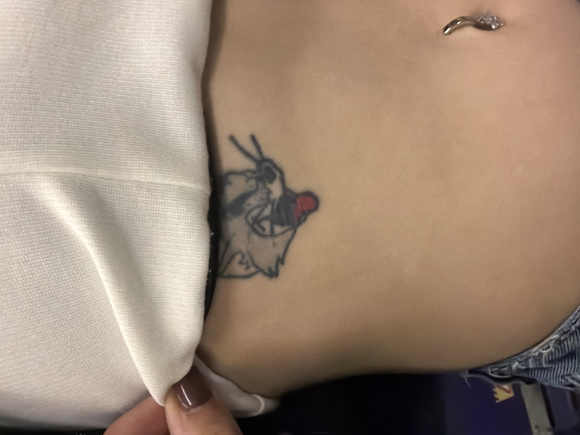 A close-up shot focuses on a section of a person's torso, where a tattoo of what appears to be a cartoon character is visible on the skin near the side. The tattoo features a figure with a red nose and dark outlines. Near the top of the frame, a piece of jewelry, likely a belly button piercing, is seen. A piece of white fabric is draped partially across the body, obscuring some of the torso, and a finger with dark nail polish is gently holding the fabric. The lower portion of the image reveals the edge of what seems to be denim clothing. The environment is indistinct, providing no contextual clues related to Sofia, Bulgaria.