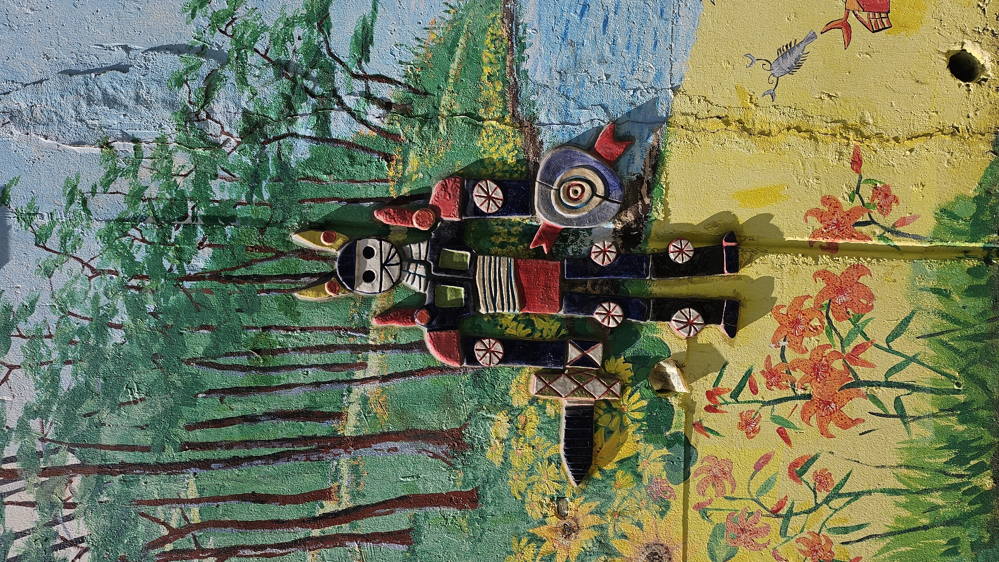 The image shows a mural on a textured wall in Ulsan, South Korea. The left side of the mural depicts a blue sky transitioning into a green forest with painted tree trunks and leaves. The right side features a yellow background with painted orange lilies. A raised, abstract, totem-like figure made of ceramic or similar material is affixed across both sections of the mural. Above the lilies, a drawing of a silver fish and an abstract red object can be seen. The background appears to be a concrete structure.
