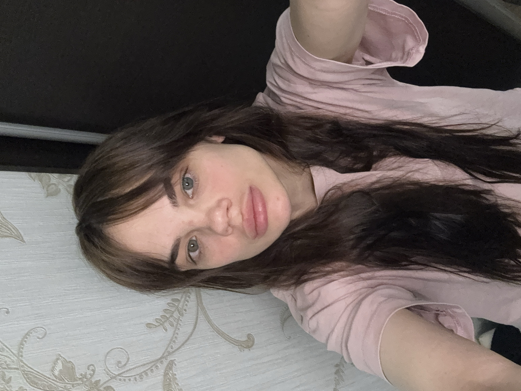 A young woman is depicted in a selfie-style photograph. She has long, dark brown hair with lighter brown highlights around her face, which is angled towards the camera. Her eyes are blue-gray, and she has full lips. She is wearing a light pink t-shirt. The background behind her consists of a patterned wallpaper with light-colored floral designs, and a dark brown or black surface above. There is no verifiable information within the image or prompt that confirms the location being in the city of Rostov-on-Don, Russia.