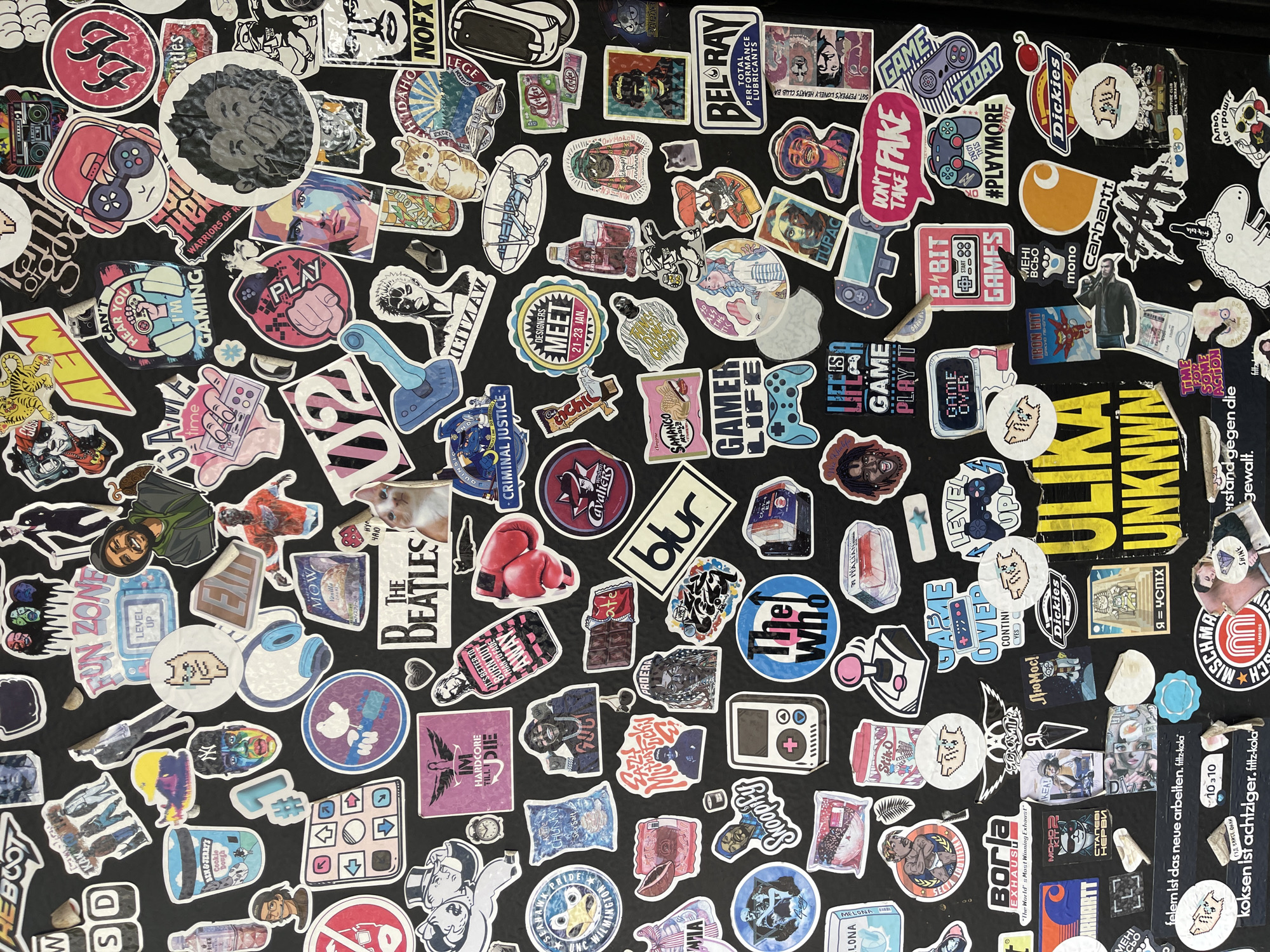 This is a vibrant, overhead shot of a densely packed collection of stickers covering a black surface. The stickers are overlapping in many areas, showing signs of wear and age, suggesting they have been accumulated over time.
The subjects on the stickers are highly diverse, encompassing various aspects of pop culture, gaming, music, and art. Prominent images include:
* **People and Characters:** Tupac Shakur, various stylized faces (some pixelated, some artistic renderings), cartoon figures, a person with dreadlocks, a boxer, a gamer character with a headset, and "The Beatles" logo.
* **Objects and Logos:** Numerous video game controllers, headphones, boxing gloves, a boombox, a skateboard, various food and drink items (e.g., Kit Kat, Ben & Jerry's, Samanco, an iced tea bottle), classic game consoles, a vinyl record, a camera, a bomb, and brand logos such as Dickies, Carhartt, NOFX, Bel-Ray, The Who, and Blur.
* **Themes:** Gaming is a strong theme, with many stickers featuring controllers, game phrases, and pixel art. Music, urban culture, and various niche interests are also represented.
There isn't a specific activity or event taking place, but rather the collective display of personal interests, fandoms, and cultural touchstones through the medium of stickers.
Notable details include the rich array of colors against the dark background. The worn appearance of some stickers adds to the sense of history and personalization.
A significant amount of text is visible across the stickers, in multiple languages:
* **English:** "GAME TODAY," "#PLVYMORE," "DON'T TAKE FAKE," "8 BIT GAMES," "LIFE IS A GAME PLAY IT," "GAME OVER," "CAN'T HEAR YOU I'M GAMING," "PLAY," "LEVEL UP," "FUN ZONE," "CRIMINAL JUSTICE," "DESIGNERS MEET 21-23 JAN," "IM HARDCORE TILL I DIE," "IT'S BETTER TO BURN OUT THAN TO FADE AWAY," "ULIKA UNKNWN," "SHINE FOR SOME ACTION," "WARRIORS OF R...", "SGT. PEPPER'S LONELY HEARTS CLUB BAND," "TUPAC," "Snoopify," "Dickies," "Carhartt," "Bel-Ray," "Borla Exhaust."
* **German:** "felern ist das neue arbeiten. filtz-kola," "koksen ist achtziger.filtz-kola."
* **Russian:** "Я = УСПІХ" (I = Success), "Альо, це грошï" (Hello, this is money), "МЕХІ ВСЬО," "МОНО."
* **Korean:** "SAMANCO."
The image captures a vibrant collage reflecting a diverse range of contemporary and classic cultural influences.