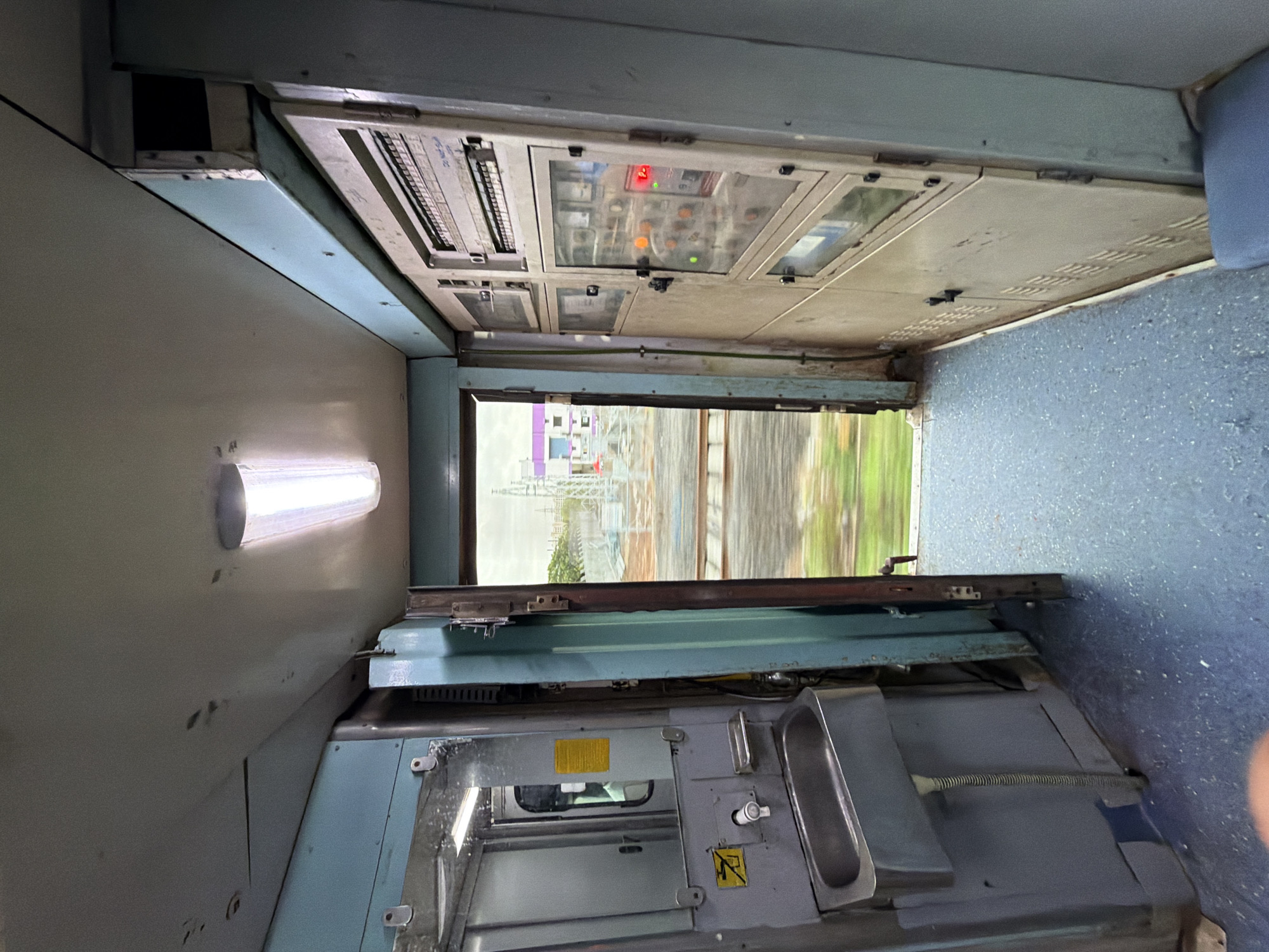 The image shows the interior of a train carriage, likely in Saphale, India. In the foreground, a blue-carpeted floor is visible. To the left, there is a grey sink with a faucet and a soap dish mounted on a blue panel. Above the sink, a mirror reflects part of the carriage. Adjacent to the sink, a yellow caution sticker is visible. To the right, a large, beige electrical panel runs along the wall, featuring multiple compartments with buttons, switches, and indicator lights, some of which are illuminated. The open doorway in the center of the image offers a view of the outside, with blurred green foliage and a track visible due to the train's motion. In the distance, buildings and a cloudy sky can be seen. The lighting inside the carriage appears to be from fluorescent tubes.