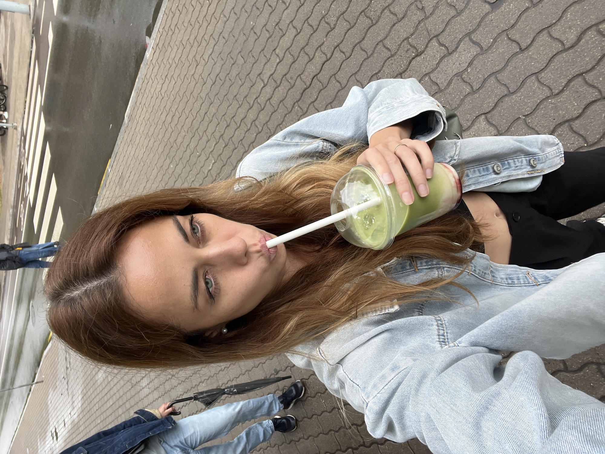 In Palanga, Lithuania, a woman with long auburn hair is captured in a close-up selfie as she sips a green beverage through a straw from a clear plastic cup. She is wearing a light blue denim jacket over a black top, and a silver ring is visible on her right hand. To her left, another person in a denim jacket and jeans, holding a closed black umbrella, walks on a patterned brick sidewalk adjacent to a wet, grey asphalt road with a white-striped pedestrian crossing. The environment appears to be an overcast day in a city setting.