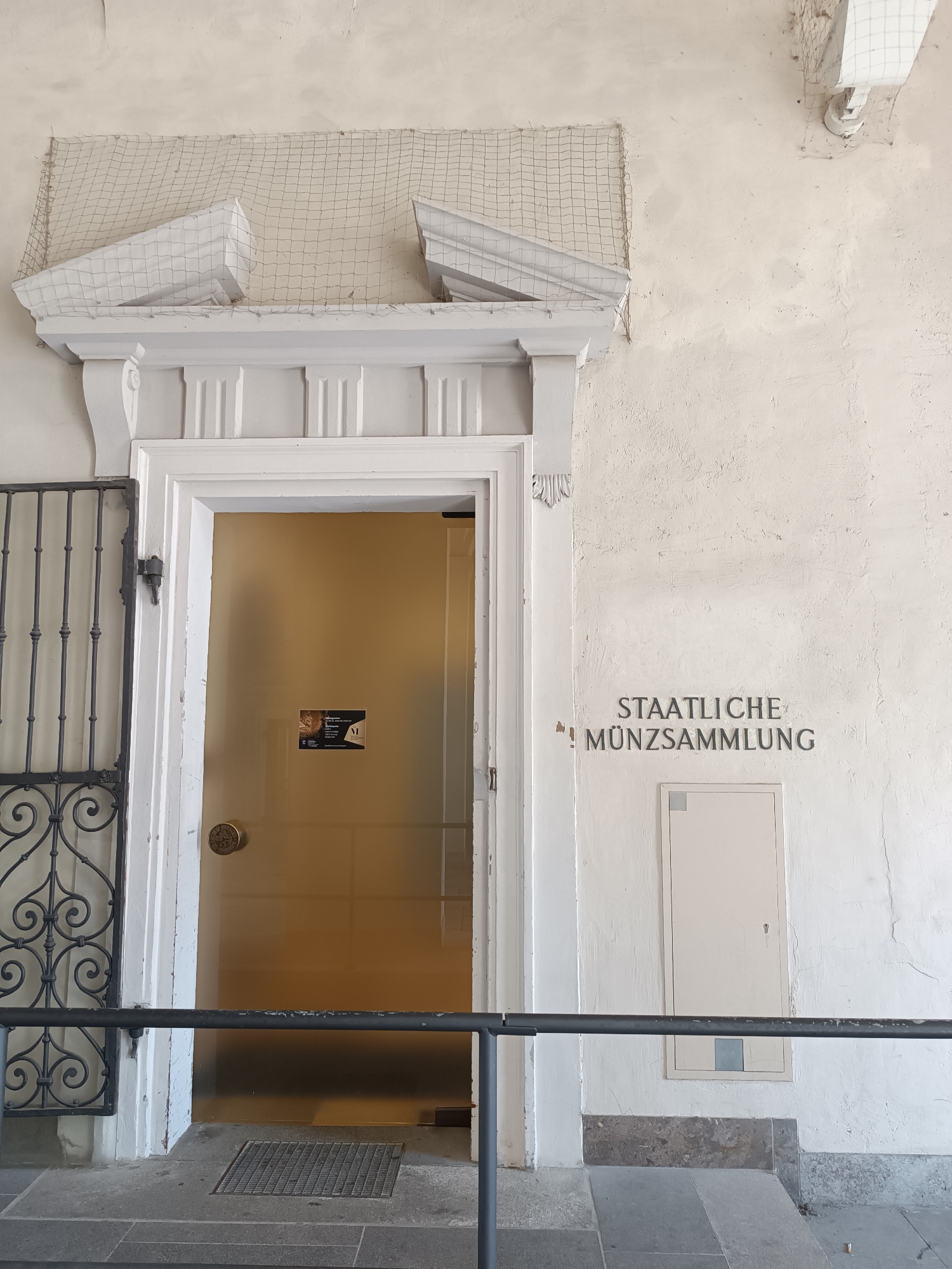 This image shows the entrance to the "Staatliche Münzsammlung" (State Coin Collection) in Munich, Germany. The entrance features a white-painted, classical style doorway with a decorative pediment. A black metal gate is visible to the left of the door, and a black metal railing runs across the lower portion of the image. The door itself is made of frosted or tinted glass, revealing a golden interior. A circular brass doorknob is present, and a small sticker is affixed to the glass. To the right of the doorway, the words "STAATLICHE MÜNZSAMMLUNG" are stenciled onto the textured, cream-colored wall. Below the lettering, there is a cream-colored utility panel with a small keyhole and a grey square patch. The ground in front of the entrance is paved with stone slabs, and a metal grate is set into the pavement. The overall scene depicts an institutional entrance in an urban environment.