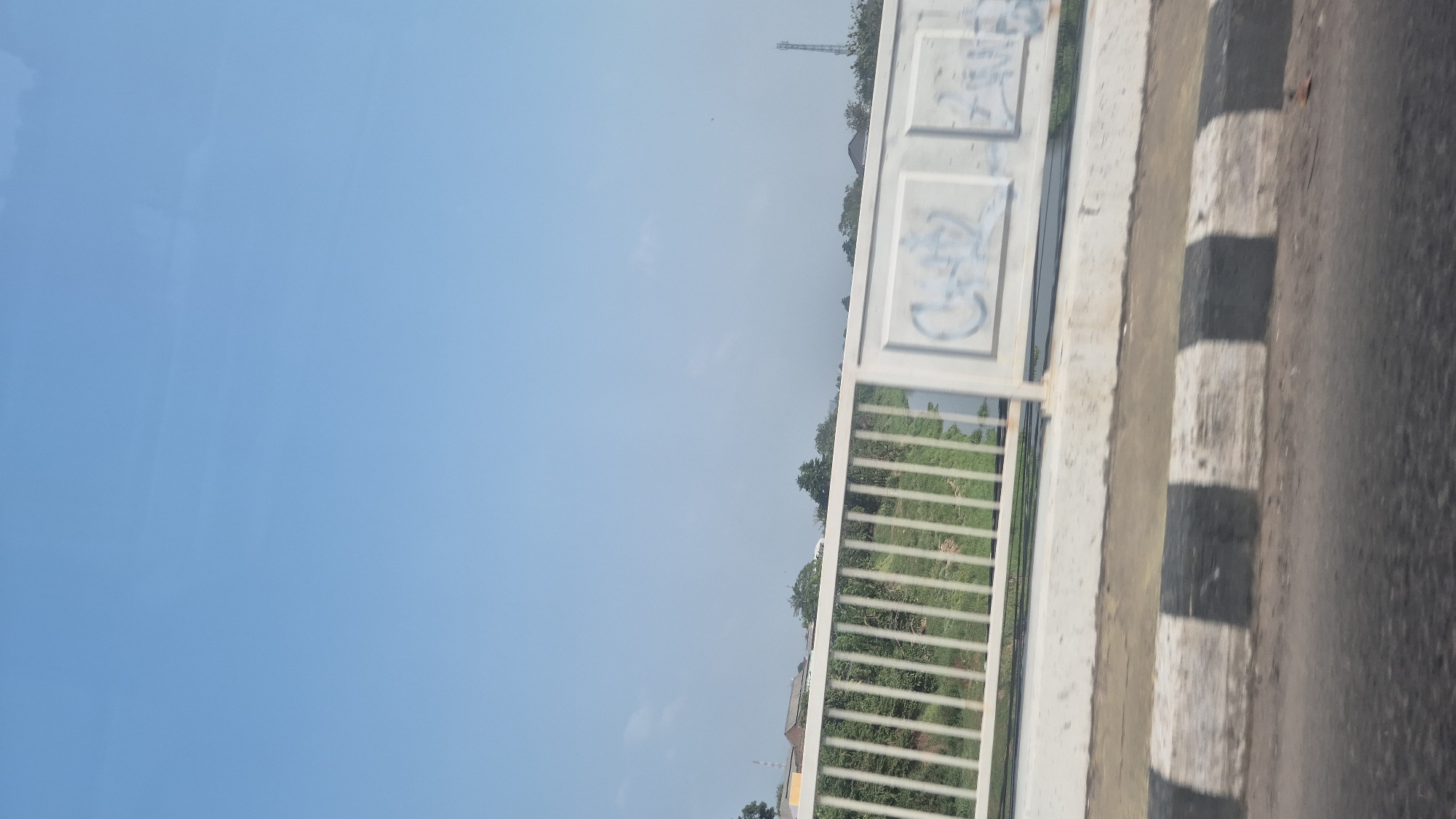 This image captures a daytime view from a bridge in Karawang, Indonesia. The sky is a clear, pale blue. In the foreground, a section of the bridge's railing is visible, composed of white vertical bars. Below the railing, a green, grassy area with some trees can be seen. On the bridge structure itself, there are panels with graffiti in white spray paint, including what appears to be stylized lettering or symbols. The edge of the bridge, demarcated by black and white painted concrete barriers, is also visible. Beyond the bridge, distant buildings and trees are present, and a telecommunications tower stands on the horizon to the right. The overall scene depicts a typical urban or suburban landscape in Indonesia, seen from an elevated perspective.
