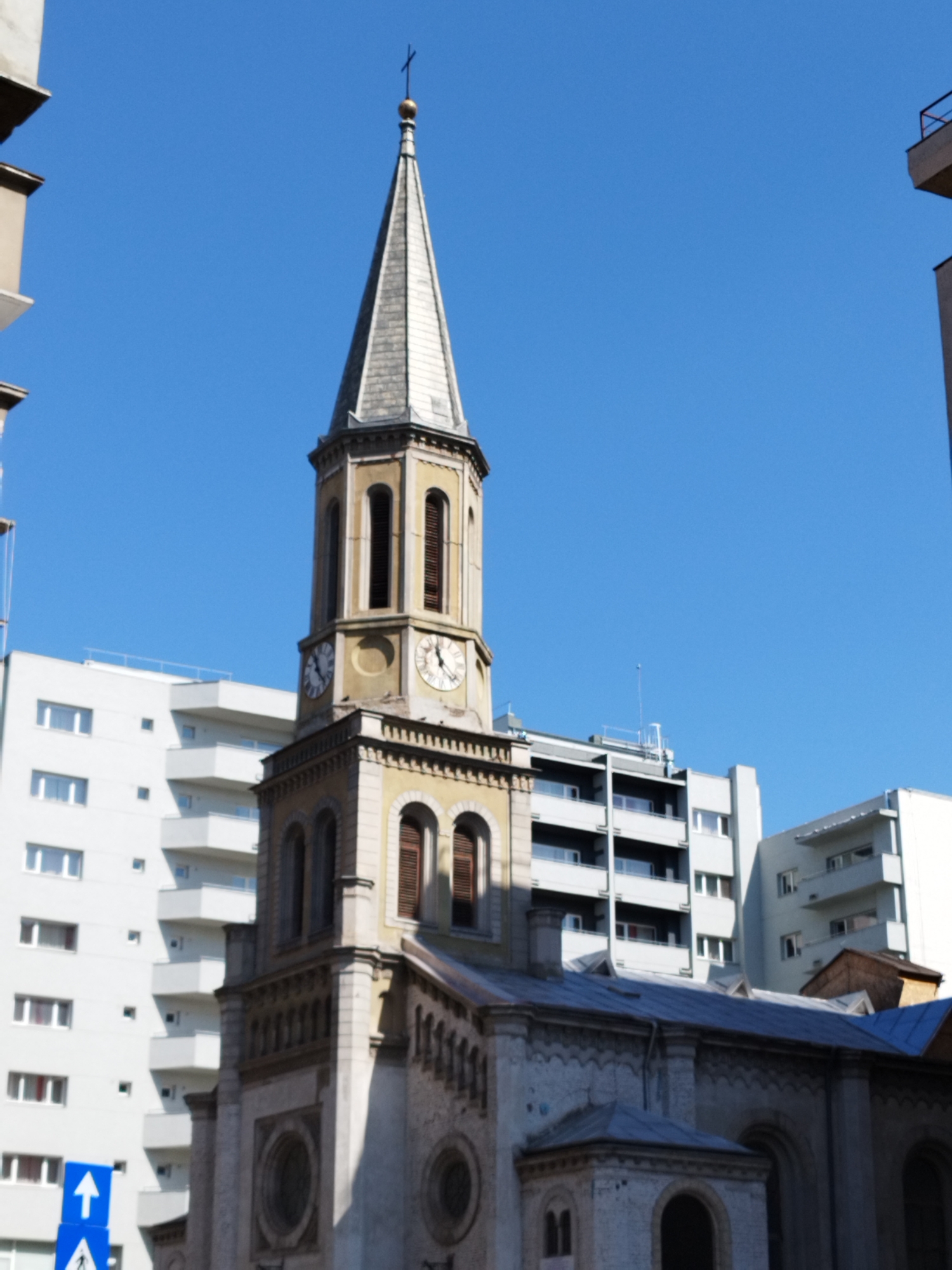 The image shows a church tower with a cross on top, set against a clear blue sky. The tower is made of stone and has arched windows. To the left and behind the church, there are modern apartment buildings with white facades and multiple balconies. A blue traffic sign with a white arrow pointing upwards and a pedestrian symbol is visible in the lower left corner of the image. The scene appears to be in an urban environment. Based on the architectural style of the church tower and the presence of modern apartment buildings, the location is likely in Bucharest, Romania, a city known for its mix of historical and contemporary architecture.