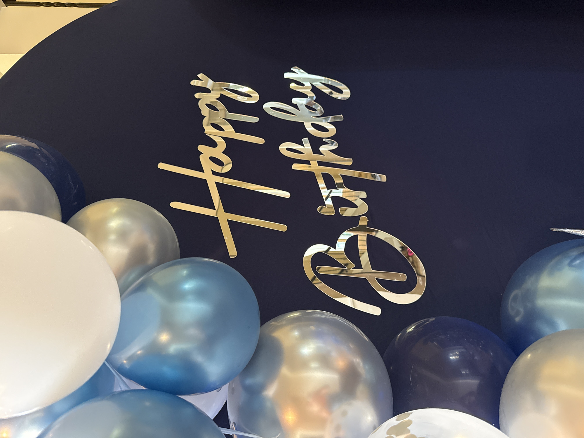 A close-up, eye-level, medium shot captures a dark navy blue backdrop with a collection of metallic balloons in white, light blue, and silver. A gold script decal that reads "Happy Birthday" is affixed to the backdrop. The balloons are clustered to the left and top of the frame, partially obscuring the backdrop and the decal. Some of the balloons are out of focus, while others are sharp. The lighting is bright and reflects off the metallic surfaces of the balloons and the decal. There is no indication of the city of Vancouver, United States of America, in the image.