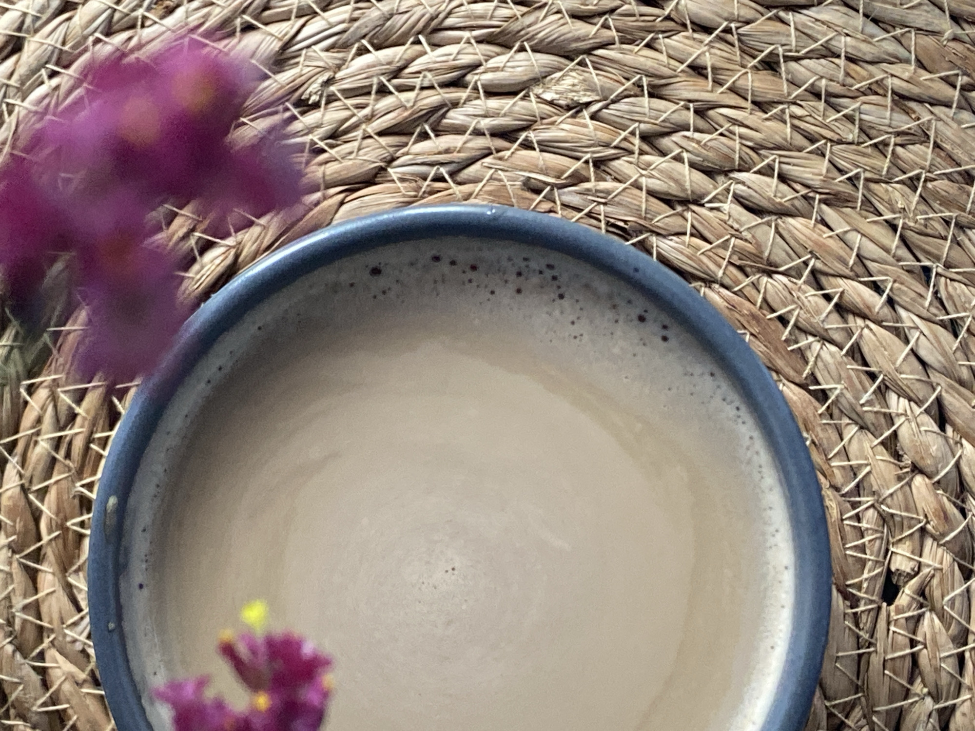 A close-up overhead shot shows a mug of light brown liquid, possibly coffee or tea, with a frothy surface, resting on a woven straw placemat. Two out-of-focus purple flowers with yellow centers are partially visible, one in the upper left of the frame and another to the right of the mug. The mug has a dark blue rim. The background is a textured straw mat with a crisscross pattern. There are no visible people or specific locations identifiable in the image.