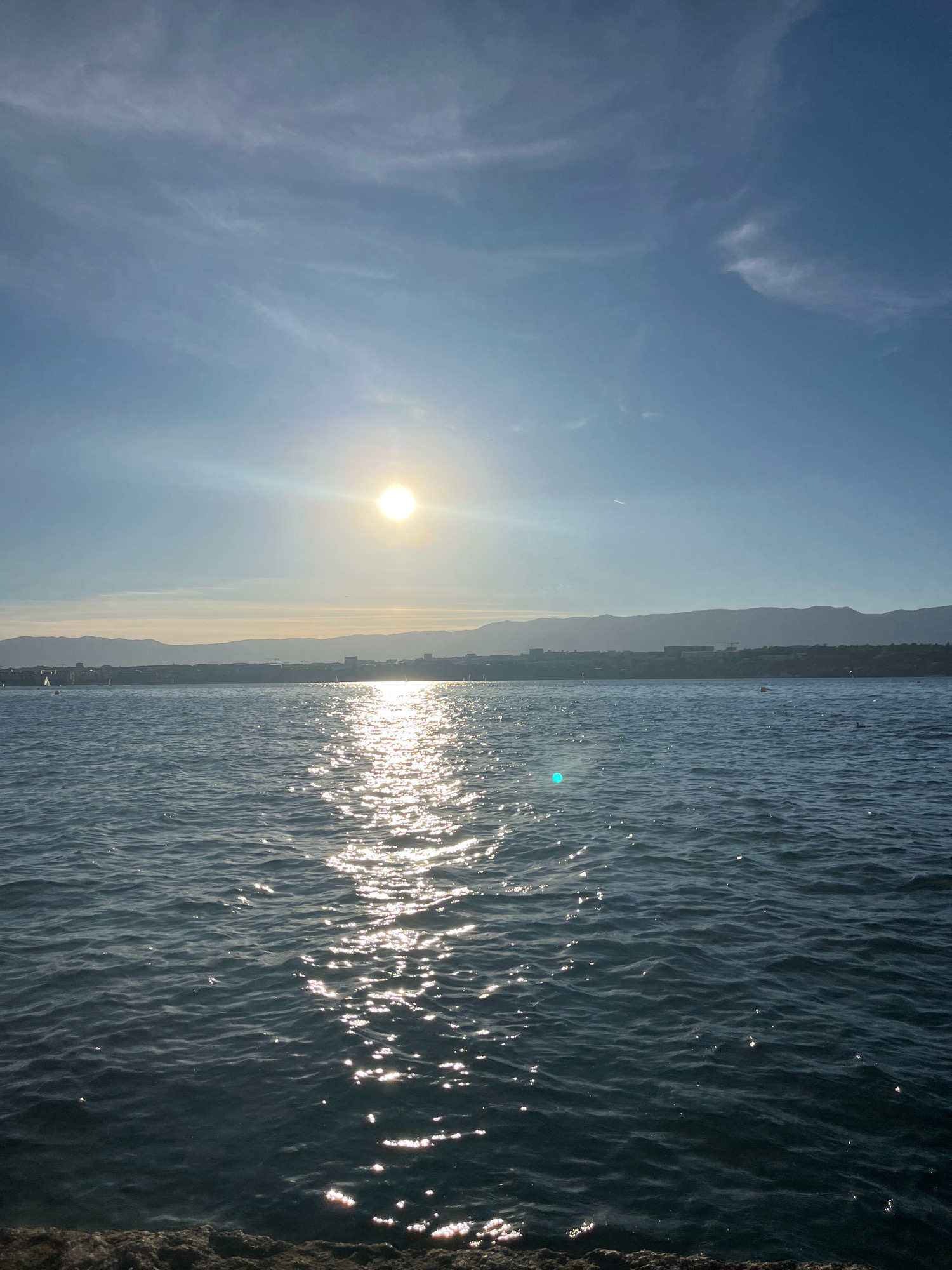 The image depicts a sunlit body of water with a distant cityscape and mountains on the horizon. The sun is positioned high in the sky, casting a bright reflection on the water's surface, creating a shimmering path of light. The water appears to be a lake, with gentle waves. Along the distant shoreline, a city skyline is visible, characterized by buildings of varying heights and some construction cranes. Further in the background, a range of mountains forms a silhouette against the sky. The sky itself is blue with scattered wispy clouds. In the foreground, a rocky edge or embankment is visible, suggesting the vantage point is from land or a structure adjacent to the water. The scene conveys a sense of daytime tranquility. The location is identified as Cologny, Switzerland, specifically showing the view across Lake Geneva towards the city of Geneva and the Salève mountain.