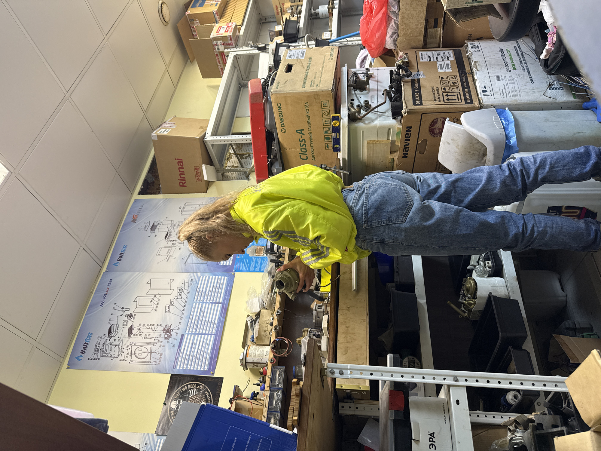 The image depicts a person with long blonde hair, wearing a neon yellow jacket and jeans, examining a mechanical component. They are standing in a cluttered workshop or storage room with shelves full of boxes and parts. The workshop appears to be in Perm, Russia based on the presence of Russian text on boxes and posters on the wall. The posters contain technical diagrams and branding for "BaltGaz" and "NEVALUX 8224." There are also boxes labeled "DAESUNG" and "NAVIEN," possibly indicating boiler or heating system components. A box labeled "Rinnai" is visible. The person is focused on the component they are holding, suggesting they may be repairing or inspecting it. The environment is cramped and filled with various tools, boxes, and mechanical parts.