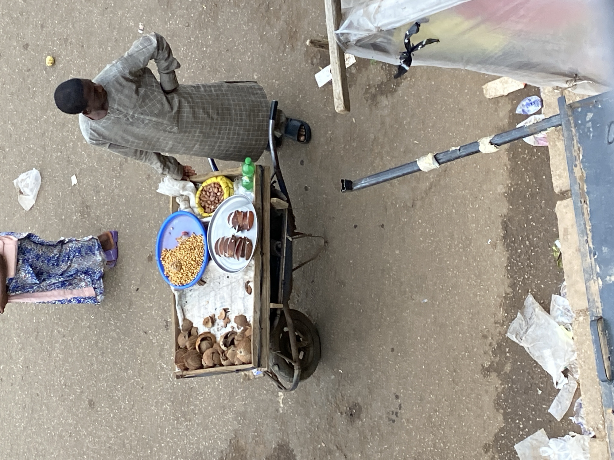 This overhead image depicts a street vending scene in what appears to be a market or roadside setting in Bukuru, Nigeria.
The main subject is a man standing beside a small, single-wheeled vendor's cart. He wears a light-colored, checkered traditional garment, possibly an agbada, and a dark cap. He is looking downwards or slightly away from the camera. His left hand rests on the cart. On the ground to the left, partially visible, is another person (likely a woman) sitting, wearing a patterned blue and white fabric and sandals.
The vendor's cart holds various items for sale. A yellow bowl contains unshelled peanuts (groundnuts). A blue bowl holds a heap of small, light brown, granular items, possibly millet or another grain. A silver plate displays several sliced, reddish-brown items, which could be kola nuts. A small green plastic bottle is also on the cart. The wooden section of the cart contains a pile of empty, brown, hard shells, strongly suggesting they are the remnants of kola nuts or similar produce.
The setting is an outdoor, paved area, likely a street or an open-air market floor, which appears somewhat dirty with scattered white plastic bags and other litter. A small, light-colored object, possibly a piece of food, lies on the ground near the man. To the right, a black metal pole extends into the frame, possibly part of a stall structure. There are also wooden planks suggesting makeshift constructions. The bright, even lighting indicates it is daytime. There is no visible text.