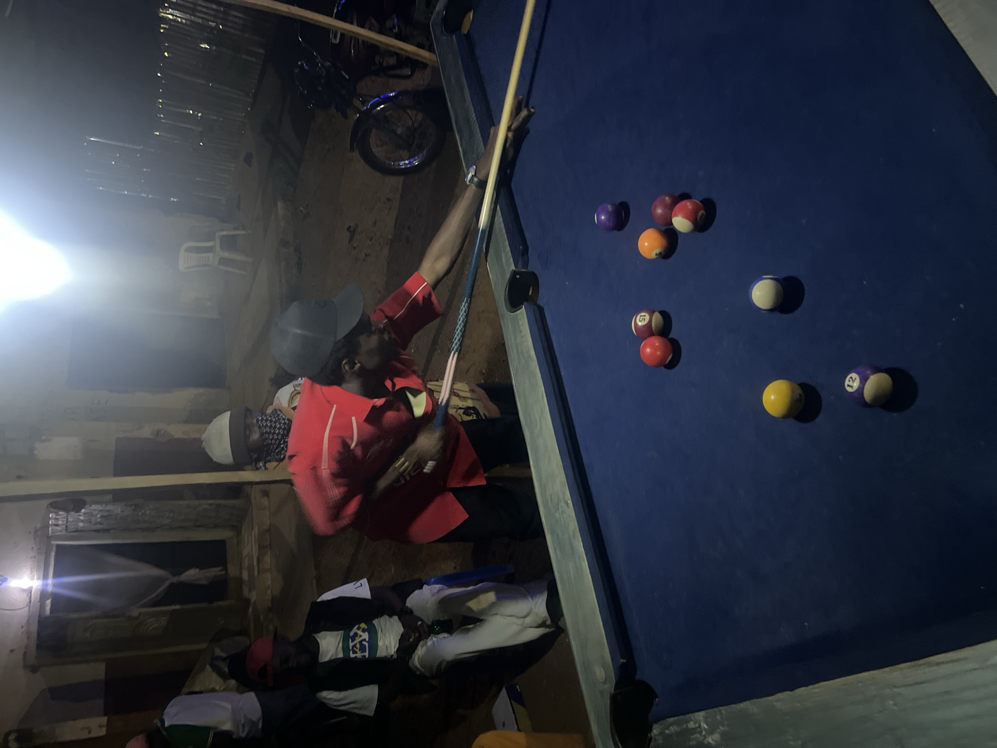 This nighttime image depicts a casual social gathering in a dimly lit, rustic setting, likely an indoor or semi-enclosed space in Gura Top, Nigeria. The central focus is a man, wearing a red long-sleeved shirt and a dark cap, actively playing pool. He is leaning over a blue-felt pool table, holding a wooden cue stick, appearing to take a shot. A watch is visible on his left wrist.
The blue pool table is populated with several colored pool balls, including solids (purple, orange, red), striped balls numbered '15' and '12', and a white cue ball.
Other individuals are present in the background. One person wearing a light-colored beanie is partially visible behind the player. Further back, another person in a white t-shirt with a graphic design and white pants is reclining or lying down next to the table, suggesting a relaxed atmosphere. The background reveals simple construction, possibly corrugated metal sheets or rough wooden planks, with visible structural beams. A white plastic chair and the wheel of a motorcycle are also discernible. A bright artificial light source, likely a bulb, creates a strong glare on the left side of the frame, confirming the nighttime setting. The overall mood is casual and engaged around the game of pool. No distinct textual information is visible other than the numbers on the pool balls.