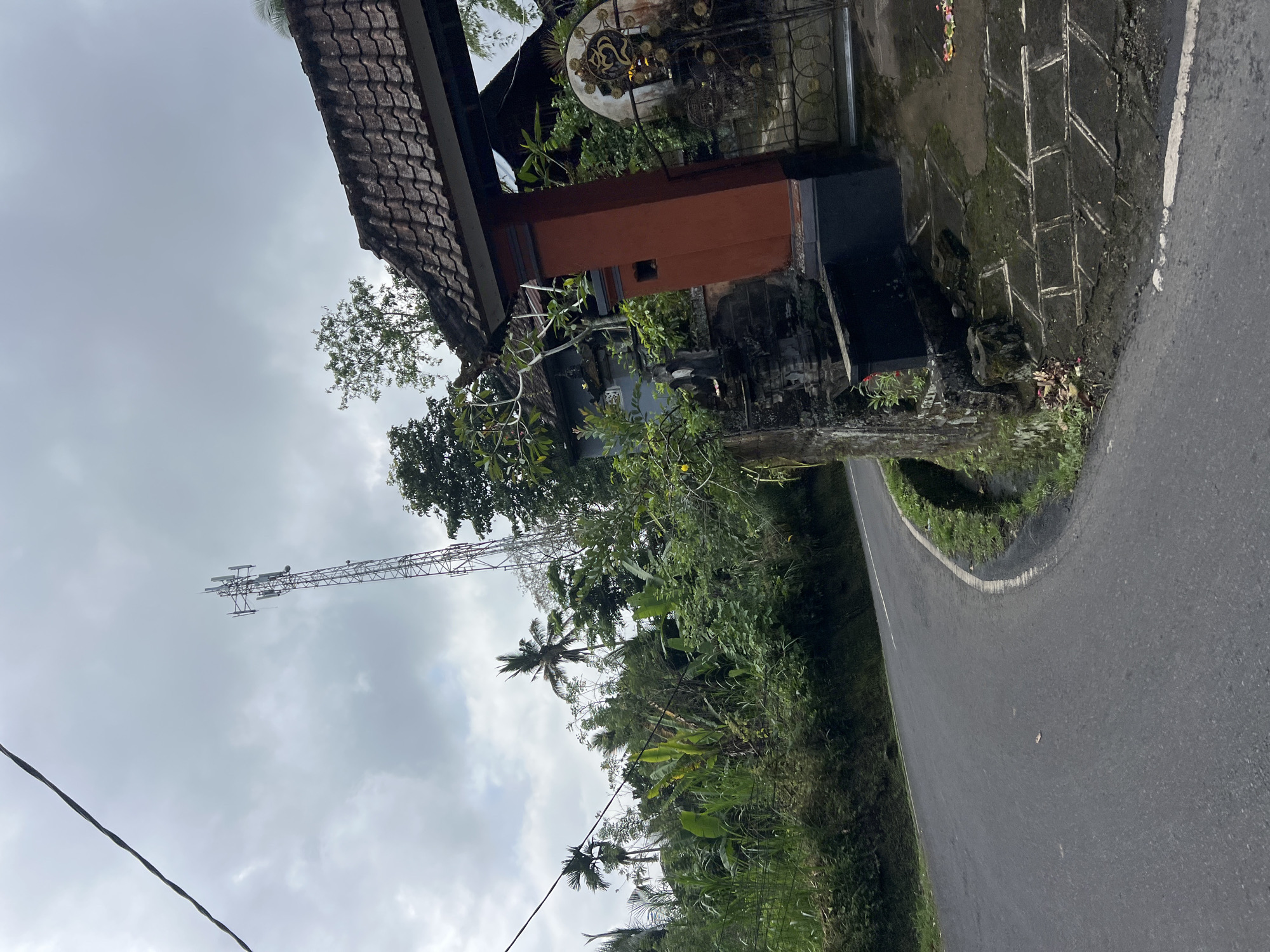 The image depicts a road winding through a lush, green landscape in Tembuku, Indonesia. A telecommunications tower stands tall in the distance, its metallic structure contrasting with the verdant foliage. On the right side of the road, a traditional Balinese gate with a tiled roof marks an entrance to a property. The gate is adorned with intricate metalwork, including an 'Om' symbol, and is flanked by ornate pillars. The surrounding vegetation is dense, with palm trees and banana plants visible, suggesting a tropical environment. The road itself is asphalt and appears to be in good condition, with a white line marking the center. The sky is overcast, with a mix of clouds and daylight. The scene is devoid of any human presence.