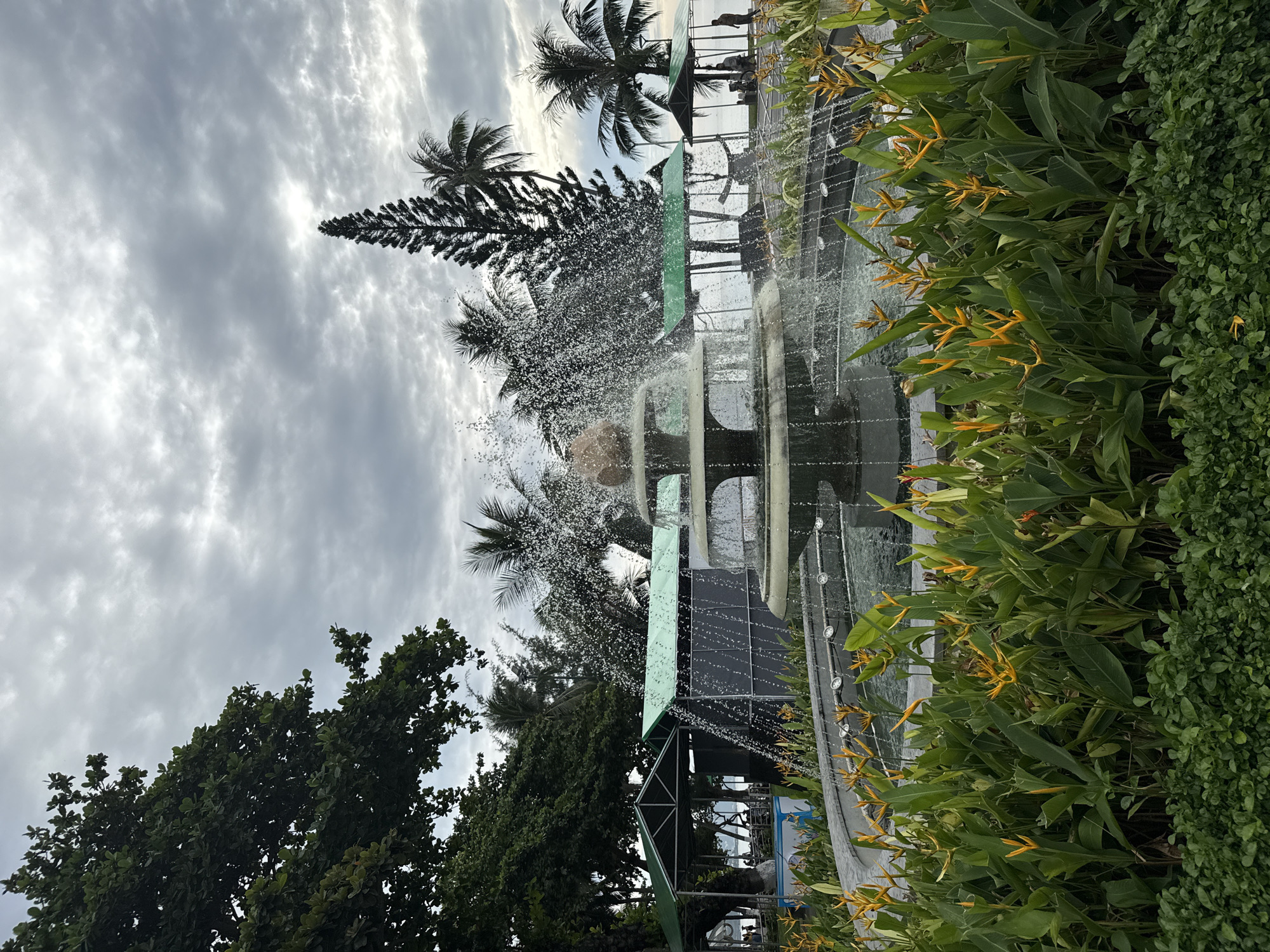 A three-tiered fountain with water cascading down is the central focus of the image, surrounded by lush green foliage and vibrant yellow flowers. Behind the fountain, several structures with green roofs are visible, with palm trees and other tropical vegetation forming a backdrop. The sky is overcast with gray clouds. In the distance, beyond the structures, a body of water, likely the sea, can be seen. A few people are present in the background, some seated and others standing, near the green-roofed structures. The overall setting appears to be a park or resort area, consistent with locations found along the coastline of Nha Trang, Vietnam.