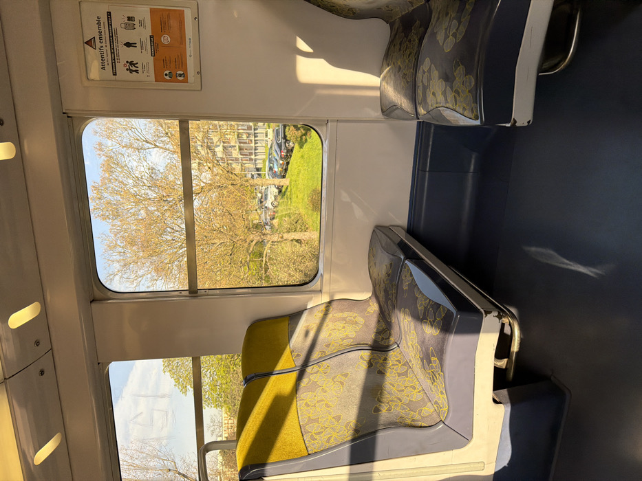 The image captures the interior of a public transport vehicle, likely a train or RER car, stopped or moving slowly in Arcueil, France. No individuals are present in the visible section of the carriage.
The interior features two empty passenger seats in the foreground, facing each other. The seat on the left is a double unit, upholstered in a grey-blue fabric with a repeating pattern of yellow, abstract, leaf-like shapes, and solid yellow headrests. The seat structure is light grey and dark blue. A single, matching seat is partially visible on the right. The floor is dark blue, while the upper walls and ceiling are white. Two oval-shaped interior lights are visible on the ceiling.
Between the two windows on the right side of the carriage, a white poster with a red triangular hazard symbol is displayed. The text "Attentifs ensemble" (Vigilant together) and "VEILLEZ A TOUTE SITUATION INHABITUELLE ?" (Pay attention to any unusual situation?) are clearly visible, accompanied by pictograms illustrating vigilance regarding luggage and suspicious behavior.
Through the windows, an exterior scene is visible under a clear blue sky with minimal cloud cover. Several deciduous trees with bare or nascent light-brown foliage stand prominently. Below the trees, a green, grassy slope descends to an area where multiple parked automobiles of various makes and colors are visible. In the background, behind the trees and parked vehicles, a multi-story building with numerous windows is discernible. Sunlight illuminates the exterior scene and casts distinct shadows within the train carriage.