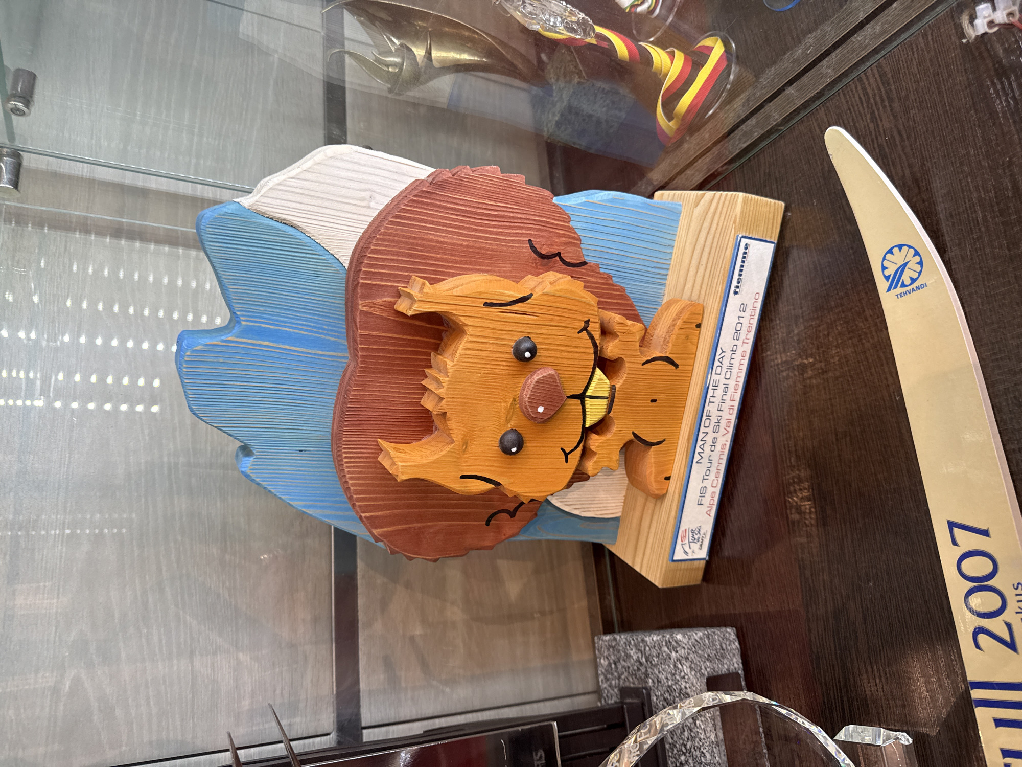 The image shows a wooden carving of a lion on a display shelf. The carving is multi-layered with different colors and textures of wood, appearing to depict a lion's head and mane on a backdrop of blue and white waves. A wooden block attached to the lion displays the words "MAN OF THE DAY," "FIS Tour de Ski Final Climb 2012," and "Alpe Cermis, Val di Fiemme Trentino," indicating that it is an award from a skiing competition. Next to the lion carving is a ski with the word "TEHVANDI" and the number "2007." Other trophies and memorabilia are partially visible in the background. Based on the information, the location is likely related to the Val di Fiemme Trentino skiing area and not Peresvet, Russia.