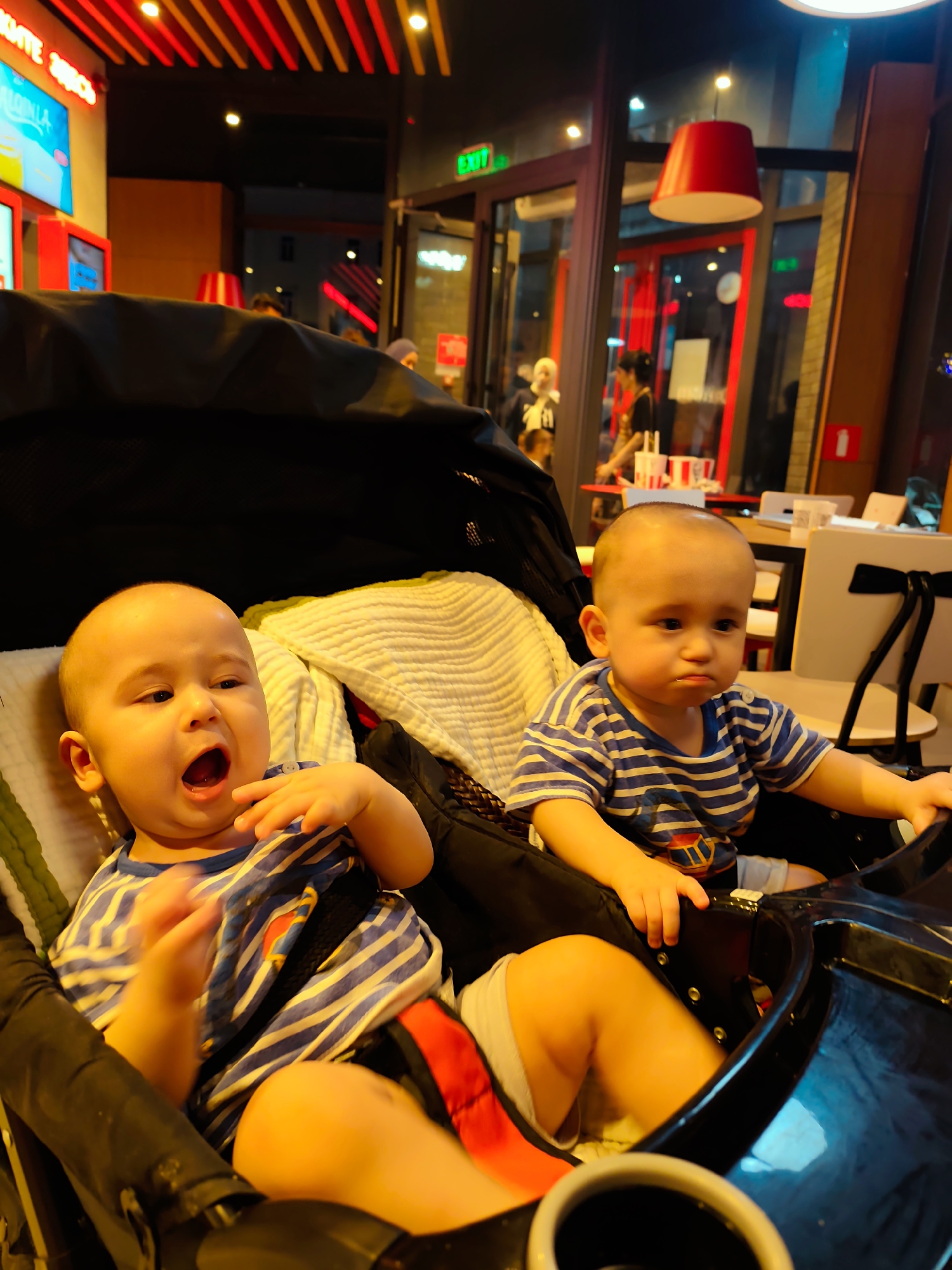 In Fergana City, Uzbekistan, two young children are seated in a stroller inside what appears to be a fast-food restaurant. The child on the left is a bald baby with an open mouth, as if yawning or speaking, and is wearing a blue and white striped t-shirt. Their left hand is raised near their mouth. The child on the right is also wearing a blue and white striped t-shirt and has a pouty expression. The stroller is black with a white blanket draped over it. In the background, a red lamp hangs from the ceiling, and through a glass door, other patrons and restaurant seating are visible. The lighting suggests it is evening.