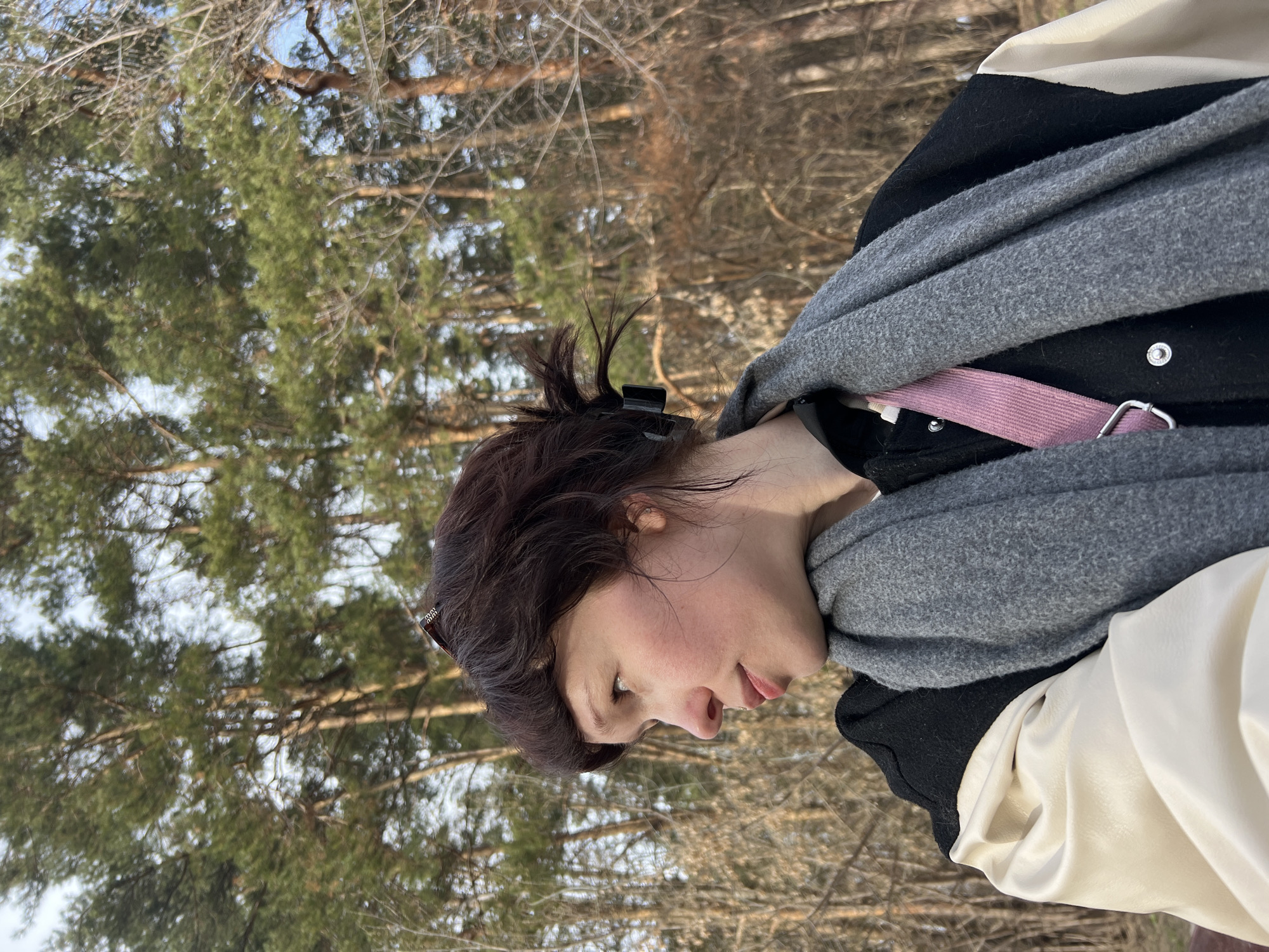 A young woman with brown hair pulled back by a black clip is looking down in a forested area. She is wearing a black and white jacket and a gray scarf. A pink strap is visible under the scarf. The background consists of pine trees and branches. The location is in a forested area and may be close to or within the city of Chaikovsky, Russia.