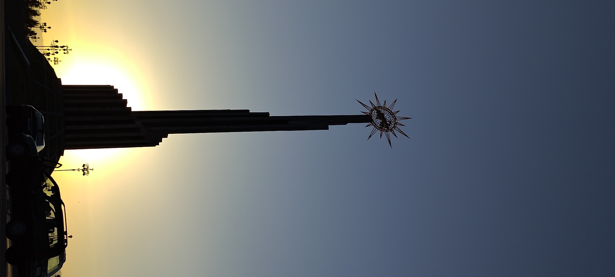 The image depicts the Arch of Ismoili Somoni in Khujand, Tajikistan, at sunset. The monumental arch, silhouetted against a gradient sky transitioning from deep blue to pale yellow, stands tall and imposing. It is topped with a distinctive starburst-like finial, intricately designed. The base of the arch is wide and tiered, creating a sense of grandeur. In the foreground, several cars are parked, casting dark shadows. Two of these vehicles, a sedan and a hatchback, are prominent. The hatchback has its passenger door ajar. Streetlights with ornate designs are visible, dotting the landscape to the left and right of the monument. The setting sun is a bright, diffused orb behind the lower section of the arch, illuminating the scene with a warm, golden light that creates a halo effect. The overall impression is one of solemnity and civic pride captured during the twilight hours.