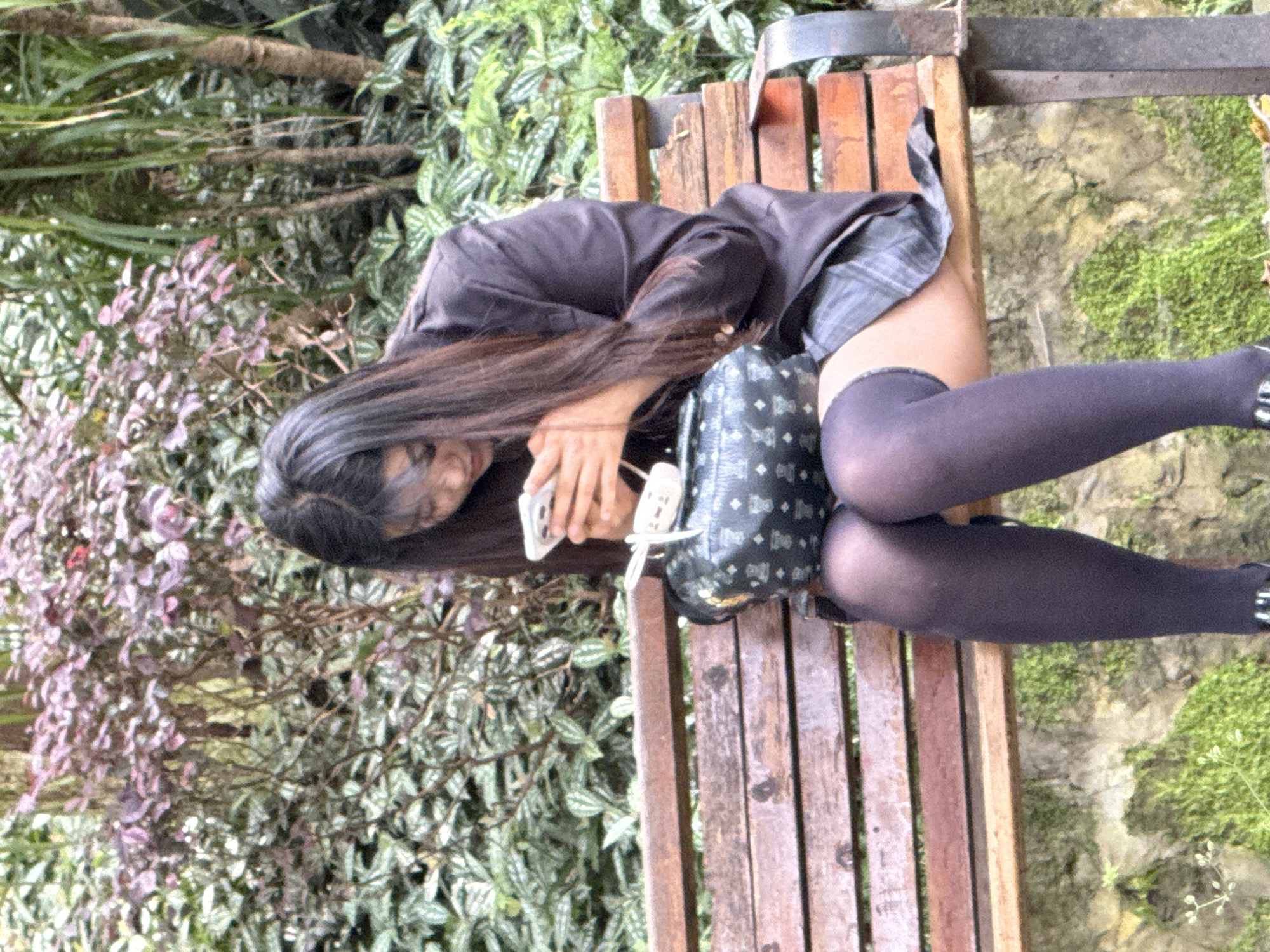 A young woman wearing a black blazer, plaid skirt, dark stockings, and studded black shoes is seated on a wooden bench in what appears to be a park or garden setting. She is holding a cell phone in her hands and appears to be looking at it. A black purse with a patterned design is positioned near her. The background consists of various plants, including ground cover, shrubs, and greenery. The location appears to be in Jiangjin, China.