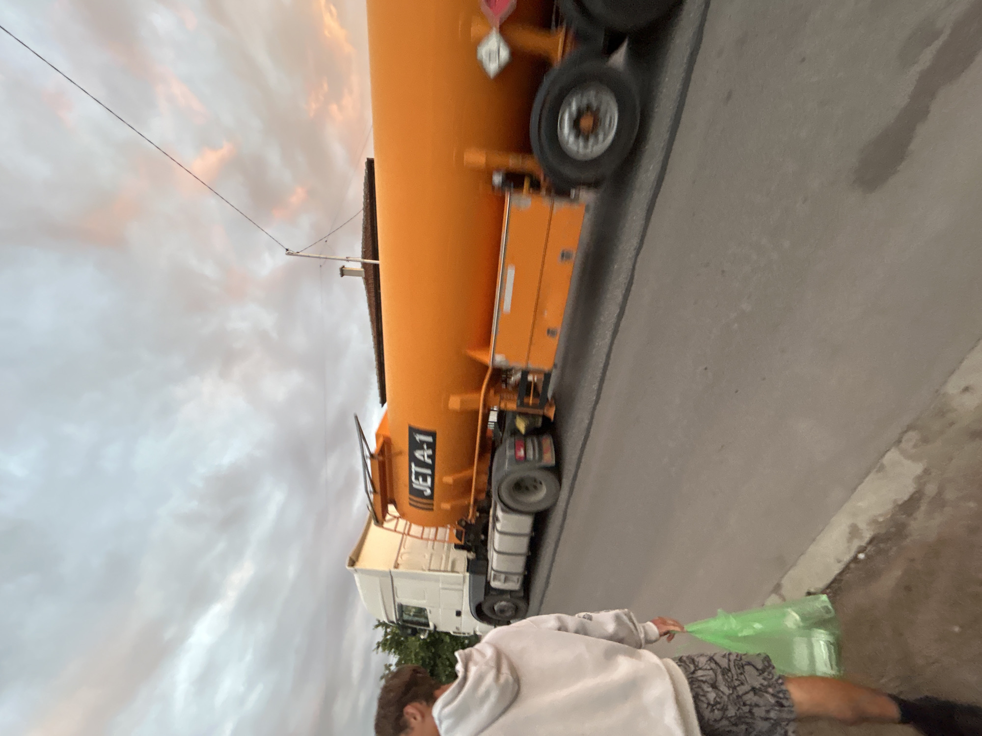A full shot shows a young person from behind, wearing a white hoodie and patterned shorts, walking on a street. The person is carrying a green plastic bag in their right hand. To the right of the person, an orange tanker truck with "JET A-1" written on its side is driving down the road. The truck has a white cab and an orange cylindrical tank. The scene appears to be in a residential or suburban area, with a building visible in the background and trees on the left. The sky is cloudy with hints of pink and orange, suggesting it may be dusk or dawn. The road is paved, and there is a concrete curb on the left side of the image.