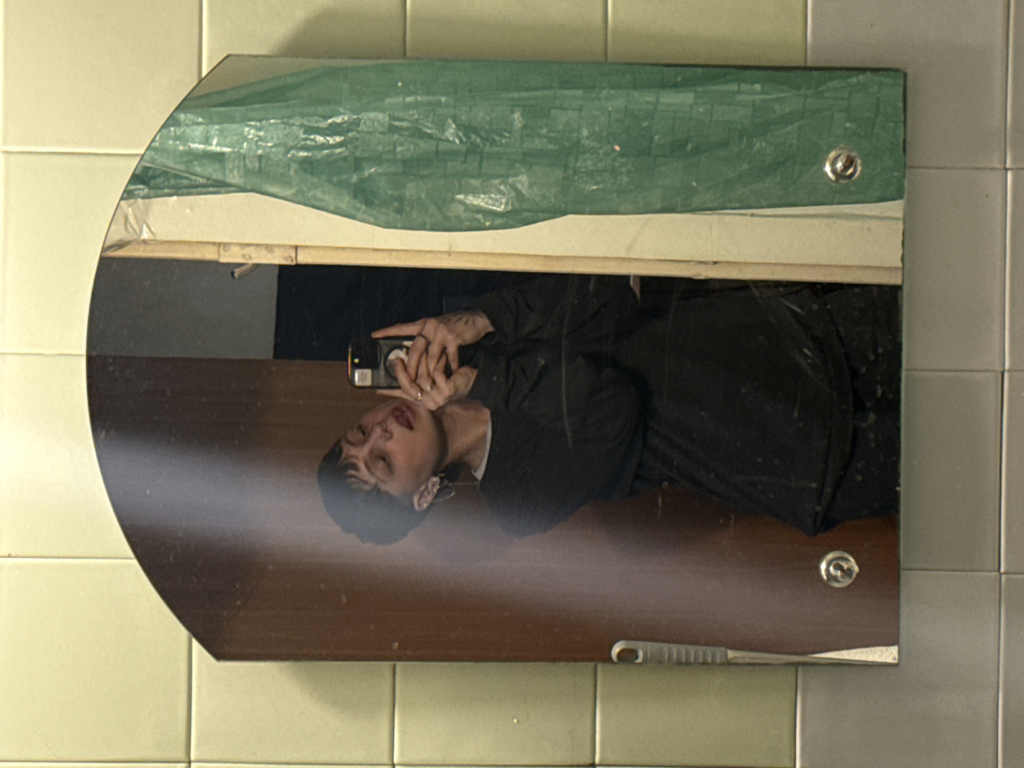 A mirror is mounted on a wall covered in light green tiles, with a person reflected in it. The mirror has a unique shape, being rectangular with a semi-circular curve on the left side. Above the mirror is a semi-transparent green sheet or tarp. The reflection in the mirror shows a person wearing a black garment, holding what appears to be a phone or camera, presumably taking a selfie.
The location is undeterminable.