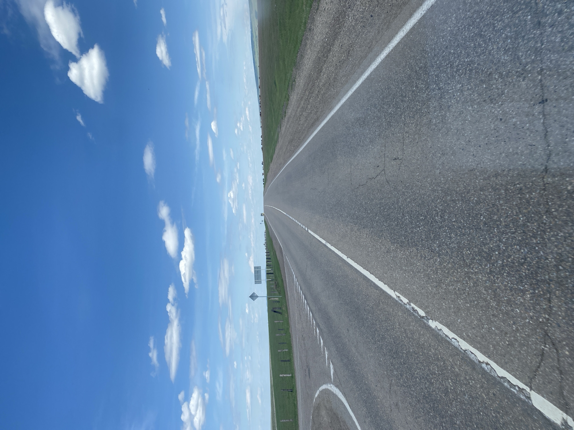 A ground-level, eye-level shot depicts a long asphalt road stretching into the distance under a partly cloudy blue sky. The road has a white center line and a solid white line along the right edge. A grassy field borders the road on both sides. A signpost is visible on the left side of the road. The scene appears to be in a rural location. The photograph has been rotated to the side. The city of Batkhai, Russia, is not clearly identifiable in the image.