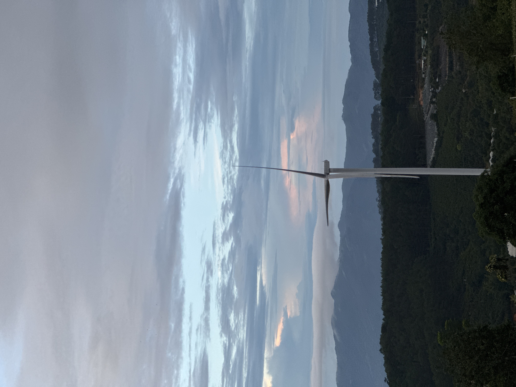 The image depicts a single wind turbine standing in front of a mountainous landscape under a cloudy sky. The turbine appears to be located near the city of Phát Chi, Vietnam, as indicated by the vegetation and terrain. The foreground shows dense green vegetation, while the background features a mountain range. The sky is filled with clouds, with hints of pink and orange suggesting either sunrise or sunset. The turbine is in focus, with one blade pointing towards the sky.