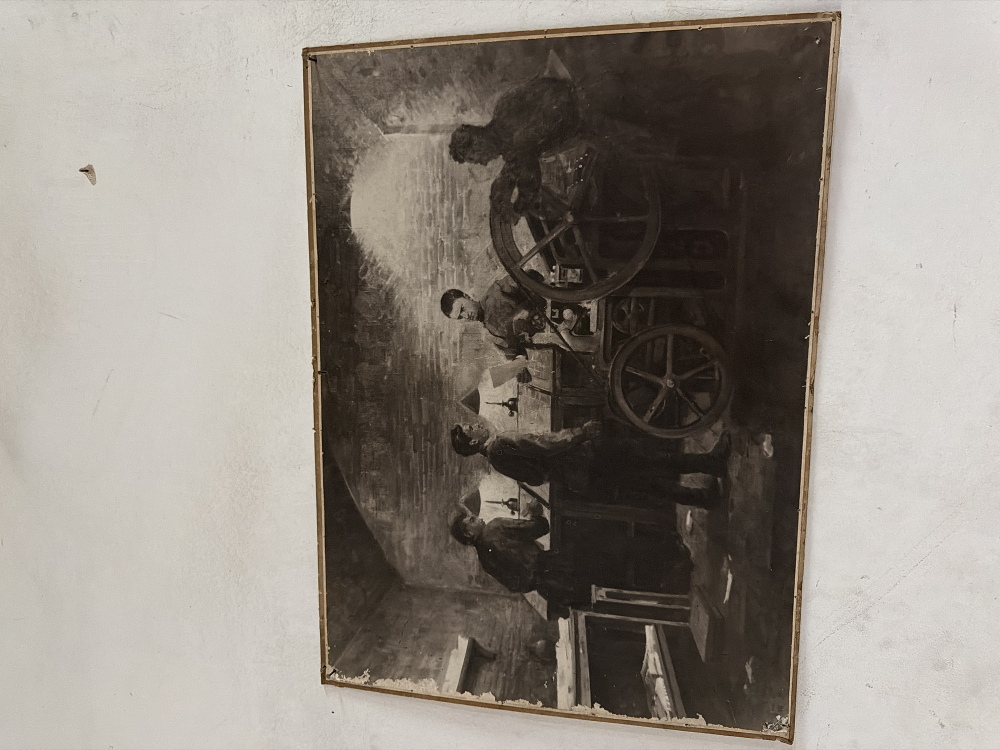 This is a black and white photograph of a painting depicting four men in what appears to be a workshop or printing facility. The painting shows the men interacting with a large printing press. The environment is dimly lit with brick walls visible in the background and what looks like a large arched opening. The painting is framed and appears to be hanging on a light-colored wall. There is a small nail or hook visible on the wall above and to the left of the painting. The painting itself shows signs of age and damage along its edges. The exact location in Tbilisi, Georgia, cannot be determined from the image alone.