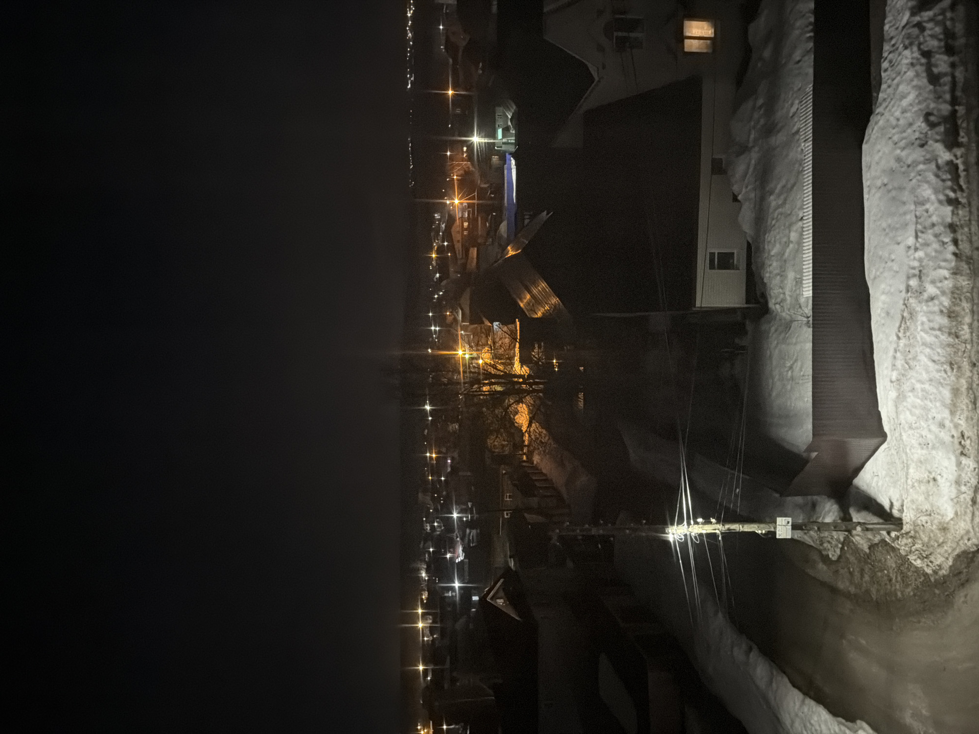 The image depicts an urban scene in Berezniki, Russia, at night. Streetlights illuminate a street lined with buildings and snowbanks. A wooden crossbeam with visible wires and attachments is visible in the foreground. The photograph appears to be taken from ground level.