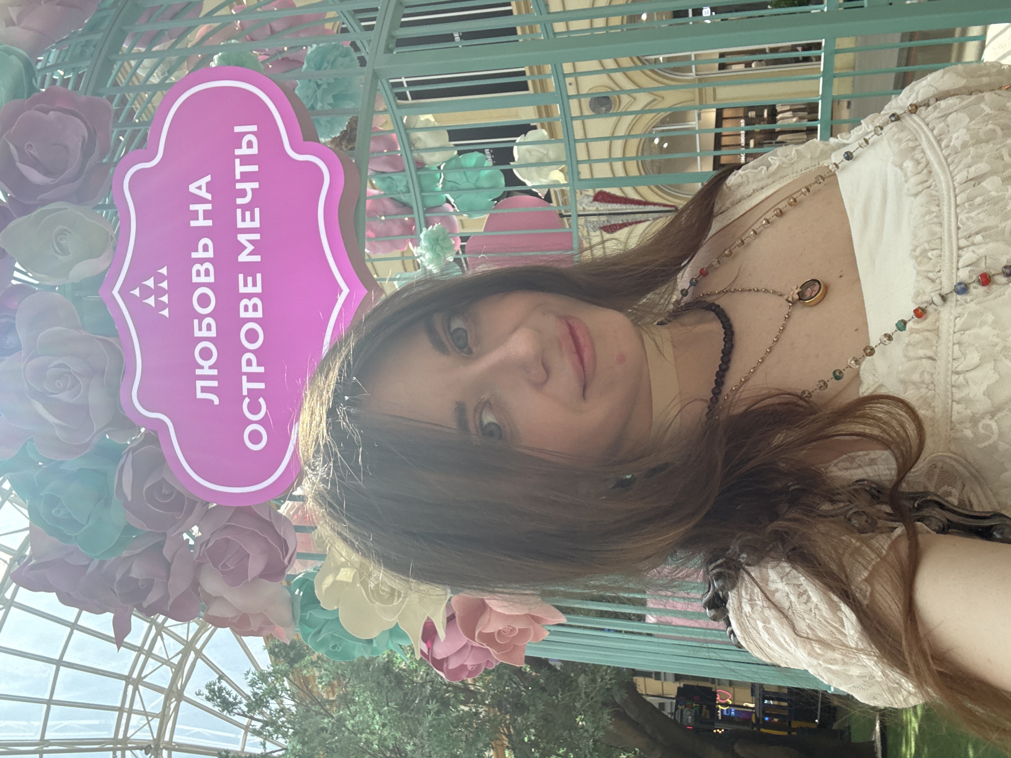 A woman takes a selfie in GUM (State Department Store) located on Red Square in Moscow, Russia. The image is rotated counterclockwise approximately 90 degrees. The woman is wearing a white lace top and multiple necklaces. To her left is a decorative structure adorned with pastel-colored paper roses and a pink sign in Russian that translates to "Love on an Island of Dreams." The interior of the GUM department store is visible in the background.