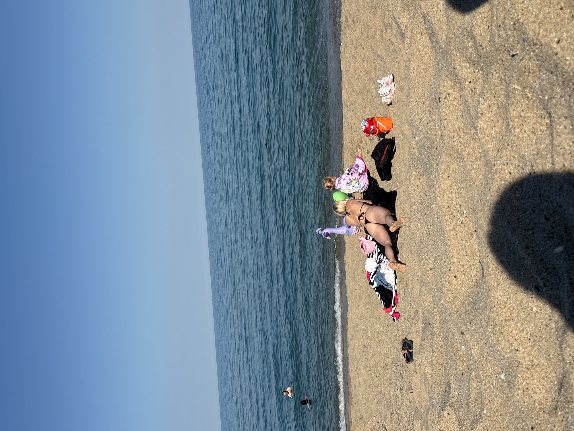 The media file captures a sun-drenched beach scene with the ocean on the left and the sandy shore on the right. The sky is a clear, pale blue. Several people are present: a woman in a bikini is lying on a towel on the sand, with another person, possibly a child, lying next to her. A woman is in the water, and another person is partially submerged. The water is a deep blue with small waves lapping at the shore.
Various objects are scattered on the sand, including a bright orange bucket, a black backpack, a pink child's shoe, and a pair of sunglasses. A colorful towel with black and white stripes is spread out on the sand, with clothing items placed on it. The sunlight casts distinct shadows on the sand, suggesting it is a sunny day, likely mid-morning or afternoon. The overall atmosphere appears relaxed and leisurely, characteristic of a beach outing. There is no visible text in the media file.