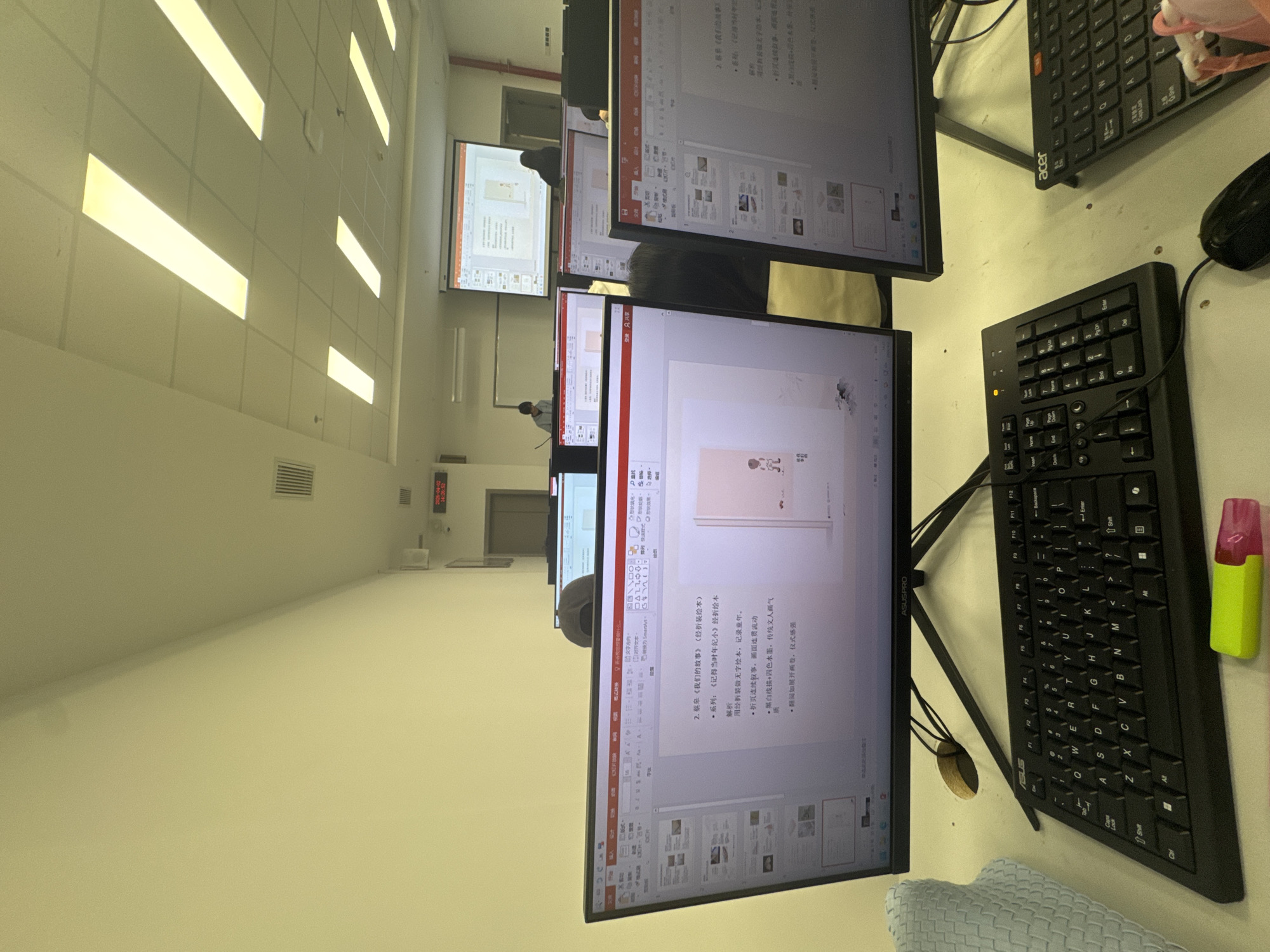 Here is a matter-of-fact description of the scene:
The image shows a classroom setting, likely a computer lab or lecture hall, in Nan'an District, China. Several computer monitors dominate the foreground, displaying what appear to be PowerPoint presentations in Chinese. Keyboards and a computer mouse are visible on the desks next to the monitors. A highlighter is also present on the desk. In the background, more computers and monitors are visible, along with a projection screen displaying similar content. People are sitting at some of the computers, presumably students or attendees of a presentation or lecture. The room has fluorescent lighting and a neutral-colored wall.