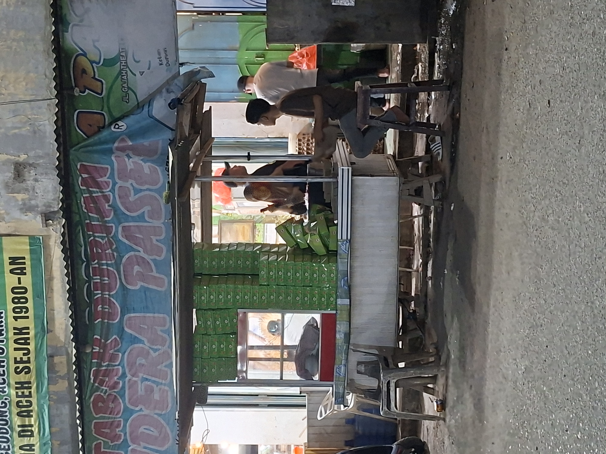 Here is a detailed description of the image:
**Scene:** A street scene in Keudegeudong, Indonesia, featuring a food stall with customers.
**People:**
* Several men are near the stall, possibly customers or workers. One man is seated on a small stool.
* People are mostly wearing casual clothing.
**Objects:**
* **Food stall:** A makeshift stall with a corrugated metal roof and signage. The signs read: "TABAK DURIAN," "DERA PASE," "IA DI ACEH SEJAK 1980-AN," and "JL. GAJAH THEATER."
* **Products:** Stacks of green boxes, possibly containing food items for sale.
* **Street:** Asphalt road with evident wear.
* **Other:** Small stools, possibly for customers.
**Actions:**
* People are interacting near the stall, likely buying food.
**Environment:** The scene appears to be a street in a commercial area. The presence of the stall suggests a local market or food vending environment.