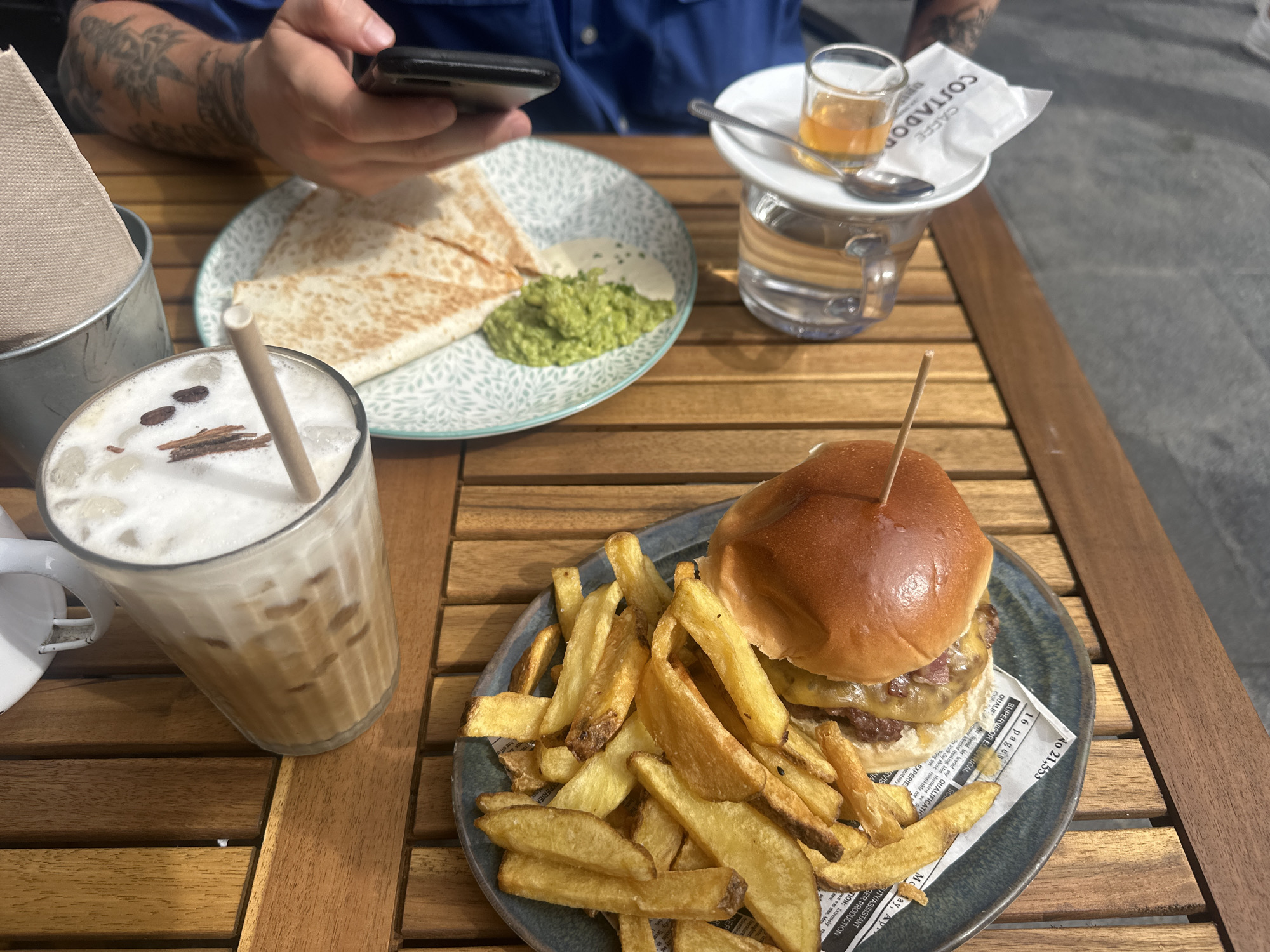 The image depicts an outdoor dining scene on a slatted wooden table, likely in Barcelona, Spain, given the "CAFFE COSTADOR" branding visible on a napkin. One individual's tattooed forearms and hands are visible, holding a black smartphone.
On the left, a tall glass contains an iced coffee with foam, ice, and a garnish of three coffee beans and several cinnamon stick pieces, accompanied by a light brown paper straw. Adjacent to it is a small silver bucket containing beige napkins. In the center, a light blue plate with a white leaf pattern holds three triangular pieces of quesadilla or flatbread, served with a dollop of white sauce and a portion of green mashed avocado or guacamole.
On the right side of the table, a small white plate supports a shot glass containing an amber liquid and a small spoon, resting on a clear glass filled with water. A white paper napkin, folded and displaying the text "CAFFE COSTADOR", is also on this small plate. In the foreground, a dark blue/grey plate holds a double-patty hamburger with melted cheese on a brioche bun, secured with a wooden skewer. Alongside the burger is a pile of thick-cut French fries, placed on a piece of patterned paper resembling newspaper. The background shows a grey, paved surface.