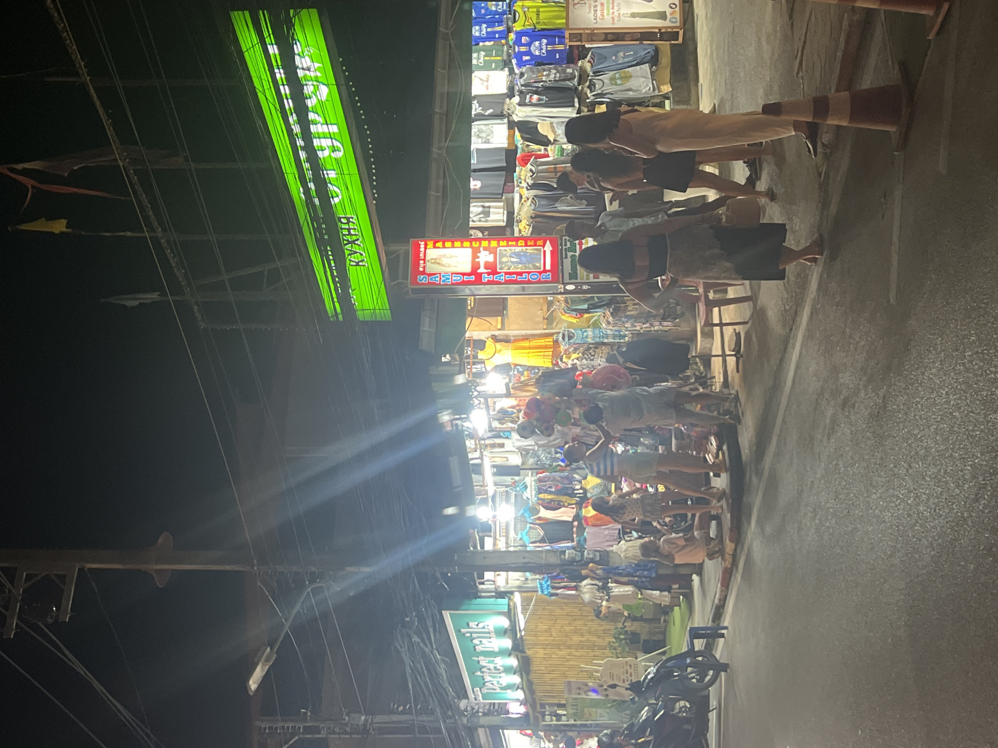 A night street scene in an urban setting shows a marketplace with several stalls and people. In the foreground, a dark asphalt road is visible with a white line marking the lane. A traffic cone with orange and white stripes stands on the right side of the road. Several people are walking and standing on the sidewalk.
The marketplace stalls display various goods, including clothing, with racks of t-shirts and dresses. A green neon sign with white text, possibly advertising a restaurant, is prominently displayed above one of the stalls. Another stall has a vertical sign that reads "Samui Tailor" with images of people. Some of the clothing items appear to be sport jerseys.
Several motorcycles are parked on the side of the street. The environment is illuminated by artificial lighting from the stalls and streetlights, creating a lively evening atmosphere. Electrical wires are visible overhead, strung between poles. A wooden fence or wall is seen behind some of the stalls. The overall scene depicts a bustling night market.