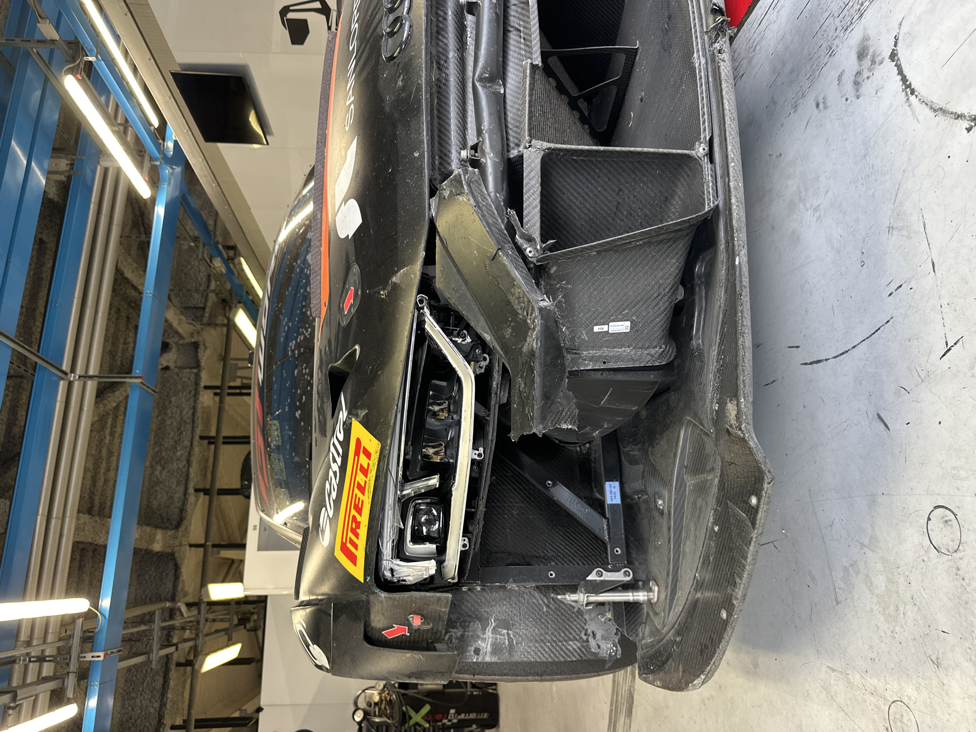 The image shows a damaged black and red sports car, likely a race car, oriented vertically in a garage-like environment. The car has visible damage to the front left, including a smashed headlight, distorted bodywork, and exposed carbon fiber structure. A "PIRELLI" sticker is visible on the front. The location is likely Monza, Italy, given the imagery and the implied presence of a race car which are common in Monza as it is home to the Autodromo Nazionale Monza, a well-known Formula One race track. The surrounding environment includes a concrete floor and industrial-style ceiling with visible lighting fixtures and ventilation systems. No people are visible in the immediate frame.