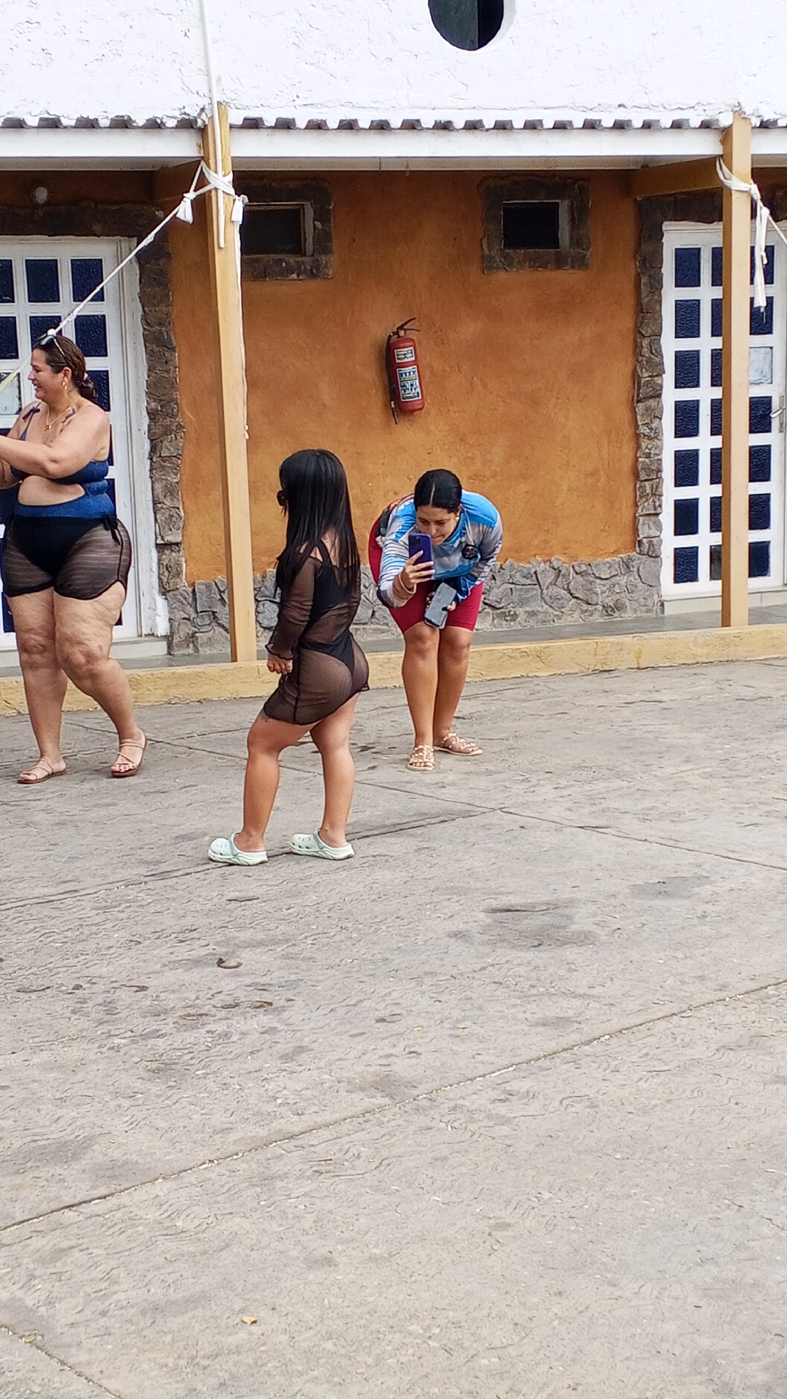 Three women are standing on a concrete surface in front of a building. The woman on the left is wearing a blue and black swimsuit with a sheer cover-up and sandals. The woman in the center is wearing a black swimsuit with a sheer dress and light-colored Crocs. The woman on the right is wearing a blue and white long-sleeved shirt, red shorts, and sandals, and is holding a cell phone up as if taking a picture. The building is painted yellow with decorative stone borders. There are three white doors with glass panes visible. A red fire extinguisher is mounted on the wall. The location is Unknown, Unknown.