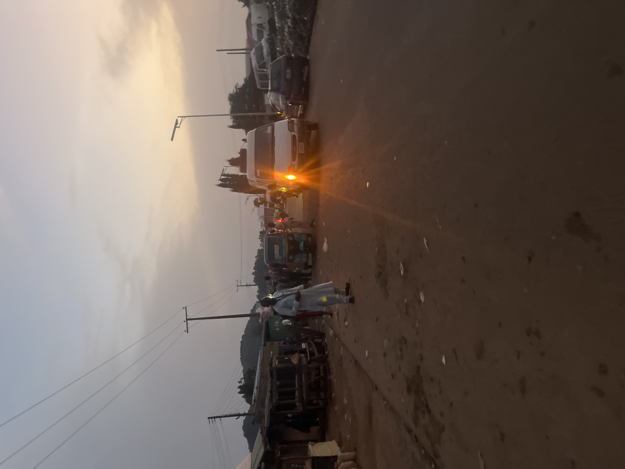 The image depicts a street scene at dusk in Gura Top, Nigeria, with a view of a dusty, unpaved road lined with informal market stalls on the left and a paved road on the right. The sky is a pale orange and grey, indicating either sunrise or sunset.
On the left side of the road, several market stalls are visible, constructed from wood and corrugated metal. Some appear to be empty, while others have goods or coverings. A few people are seen near the stalls, one individual wearing a light-colored, traditional garment is standing on the unpaved side.
The paved road on the right is active with vehicular traffic. A white minibus with its headlights on is prominent in the center, emitting a strong glare. A dark-colored car is parked or moving behind the minibus. Further down the paved road, other vehicles, including vans and what appears to be a tuk-tuk or three-wheeled auto-rickshaw, are present. The environment appears somewhat disorganized, with litter scattered on the ground. In the background, trees and hills are visible under the twilight sky. Overhead, power lines stretch across the scene, supported by utility poles.