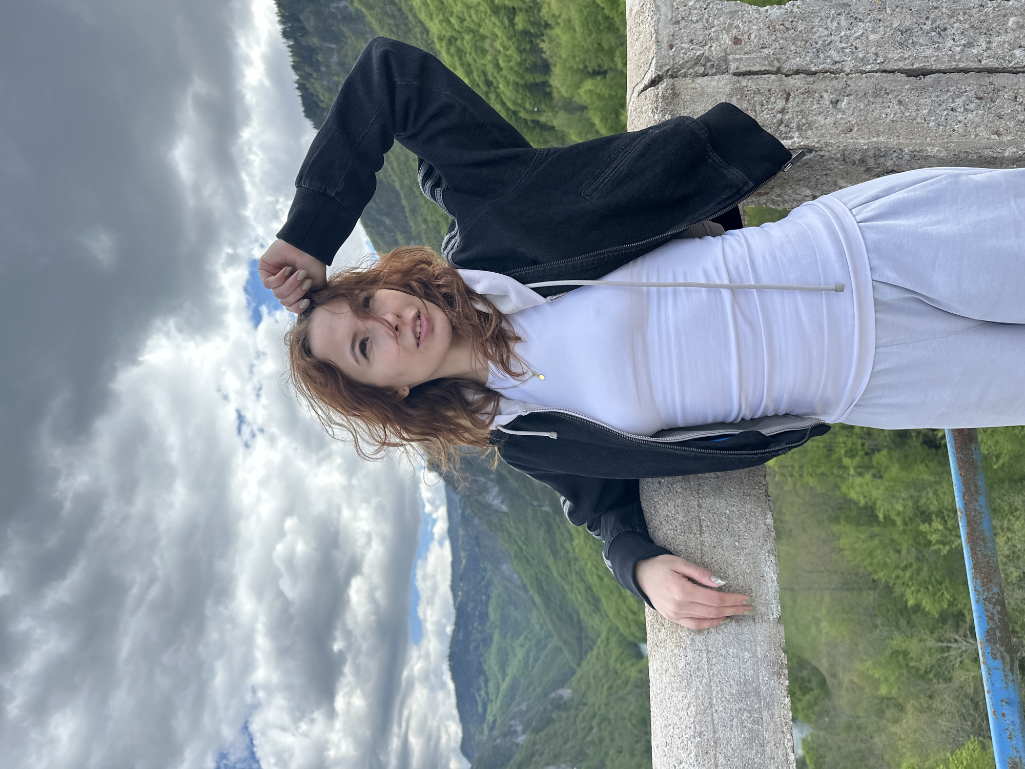 A medium shot shows a woman leaning against a concrete barrier with a landscape of mountains, trees, and sky in the background. The sky is filled with grey clouds. The woman is centered. She has red hair and is wearing a white shirt, grey sweatpants, and a black jacket. Her right arm is bent, with her hand near her head. Her left hand is resting on the barrier. The location is Đurđevića Tara, Montenegro. A blue metal barrier is visible on the right.