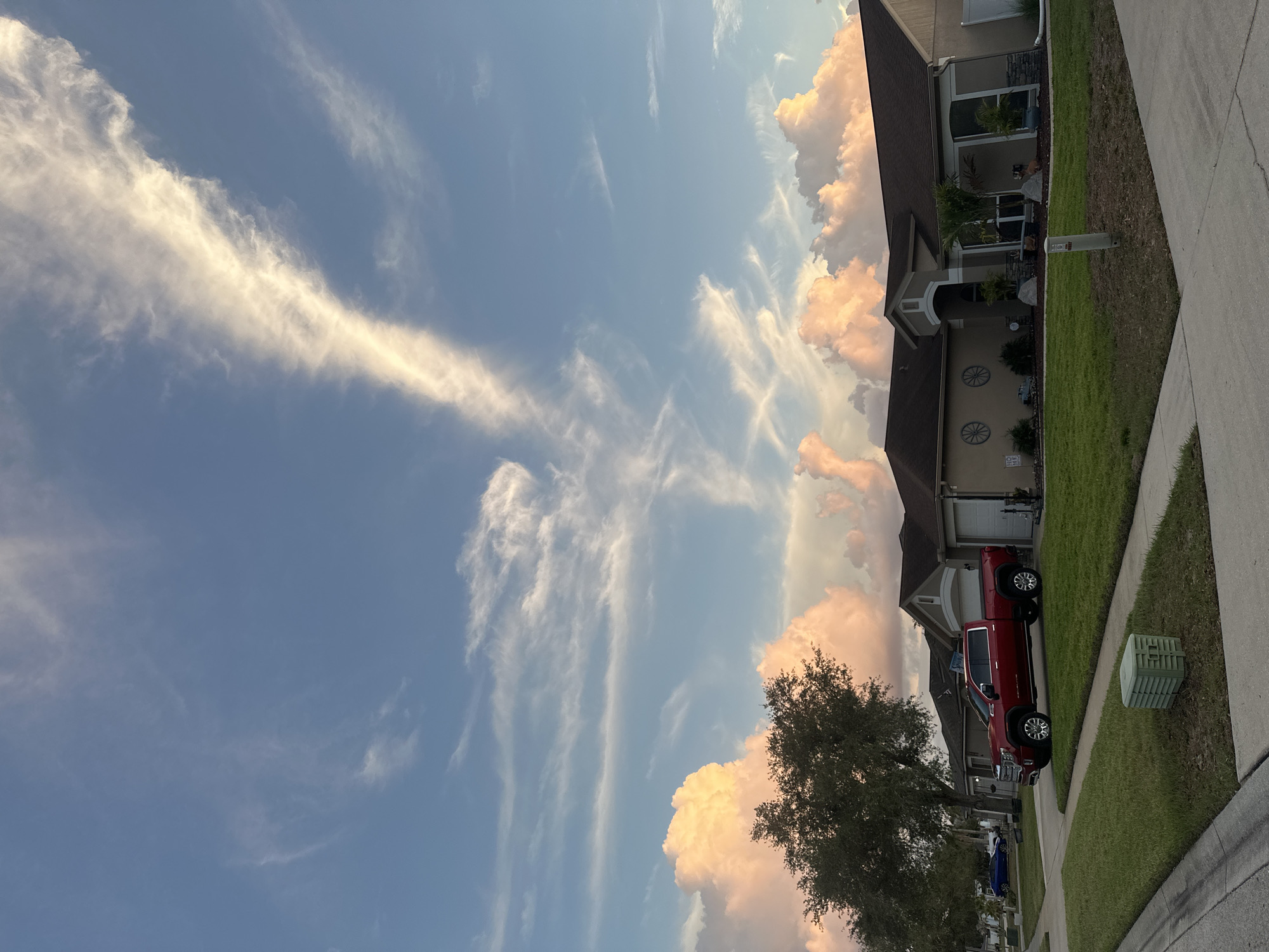 The image depicts a residential street in Riverview, Florida, United States. A red pickup truck is parked on the street in front of a beige single-story house. The house has a dark brown roof, white garage door, and decorative wagon wheels on the facade. A green utility box sits on the grassy verge between the sidewalk and the curb. The sky above is blue with wispy cirrus clouds and cumulus clouds tinged with pink hues, indicating sunrise or sunset. A tree with green foliage is visible in front of the house.
