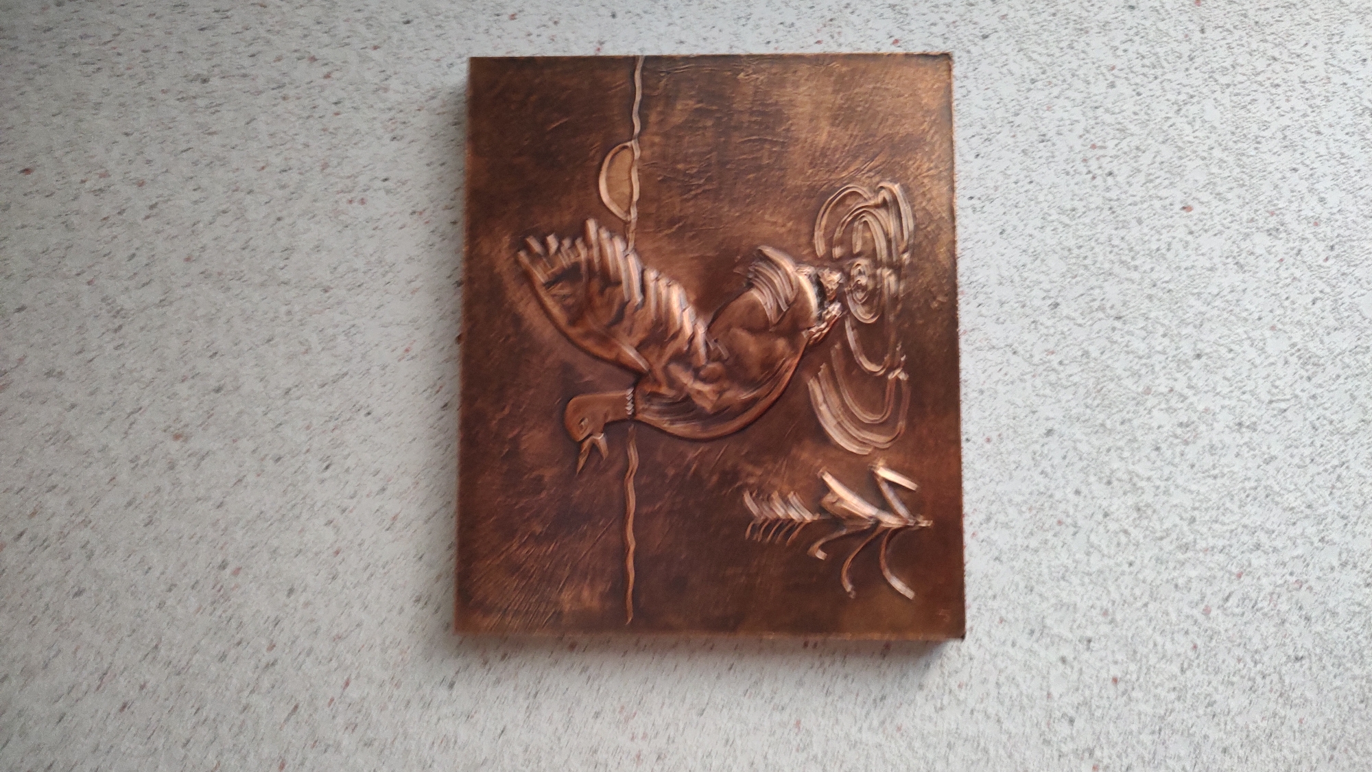 A copper relief artwork depicting a duck is mounted on a textured wall. The duck is shown with its wings spread, appearing to be either taking off or landing in water. The artwork features a horizontal line representing water, with ripples around the duck and a suggestion of reeds or grass on the left. A semi-circular shape on the right suggests a setting sun or moon. The background of the artwork has a textured, brushed effect. The wall the artwork is hung on has a speckled texture with small flecks of various colors. The image does not provide information about its location in the city of Allendorf, Germany.
