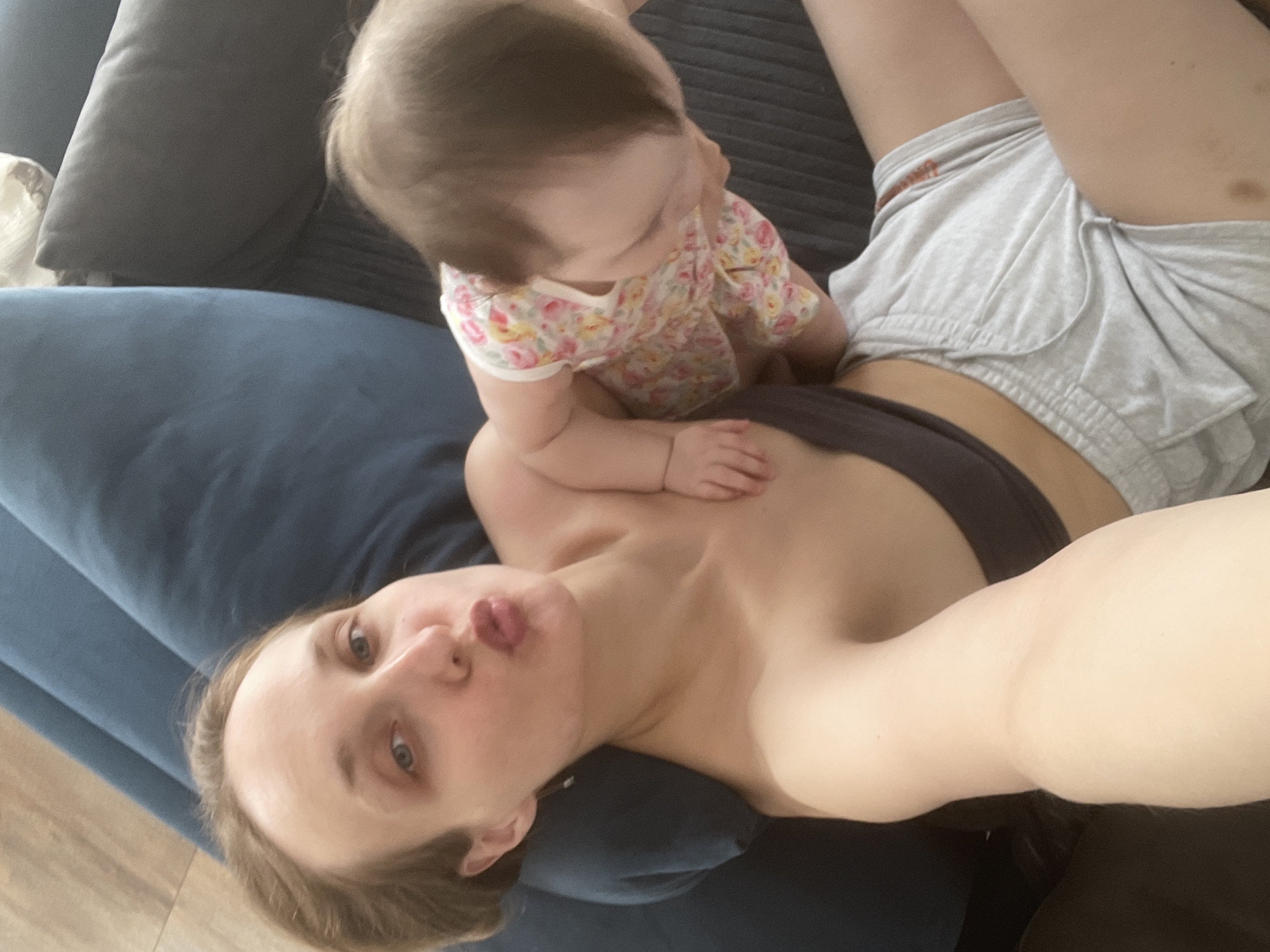 An overhead shot captures a woman and a baby sharing a tender moment on a dark blue sofa in an indoor setting, likely a home in Podshivalovo, Russia. The woman, with light hair and fair skin, lies on her back, gazing up towards the camera with slightly puckered lips, suggesting a playful or affectionate expression. She is wearing a dark sports bra or top and light grey athletic shorts. A dark cushion is visible behind her head.
The baby, with light brown hair, is positioned on the woman's chest and stomach, appearing to look down towards the woman's left side. The baby is dressed in a light-colored onesie adorned with a vibrant floral pattern of pink, yellow, and green. The woman's arm is visible supporting the baby. Part of a light-colored wooden floor is visible in the bottom left corner of the frame, providing context to the home environment. The lighting appears soft and natural, suggesting daytime. No discernible text is visible in the image. The overall scene conveys a sense of warmth, intimacy, and maternal bonding.