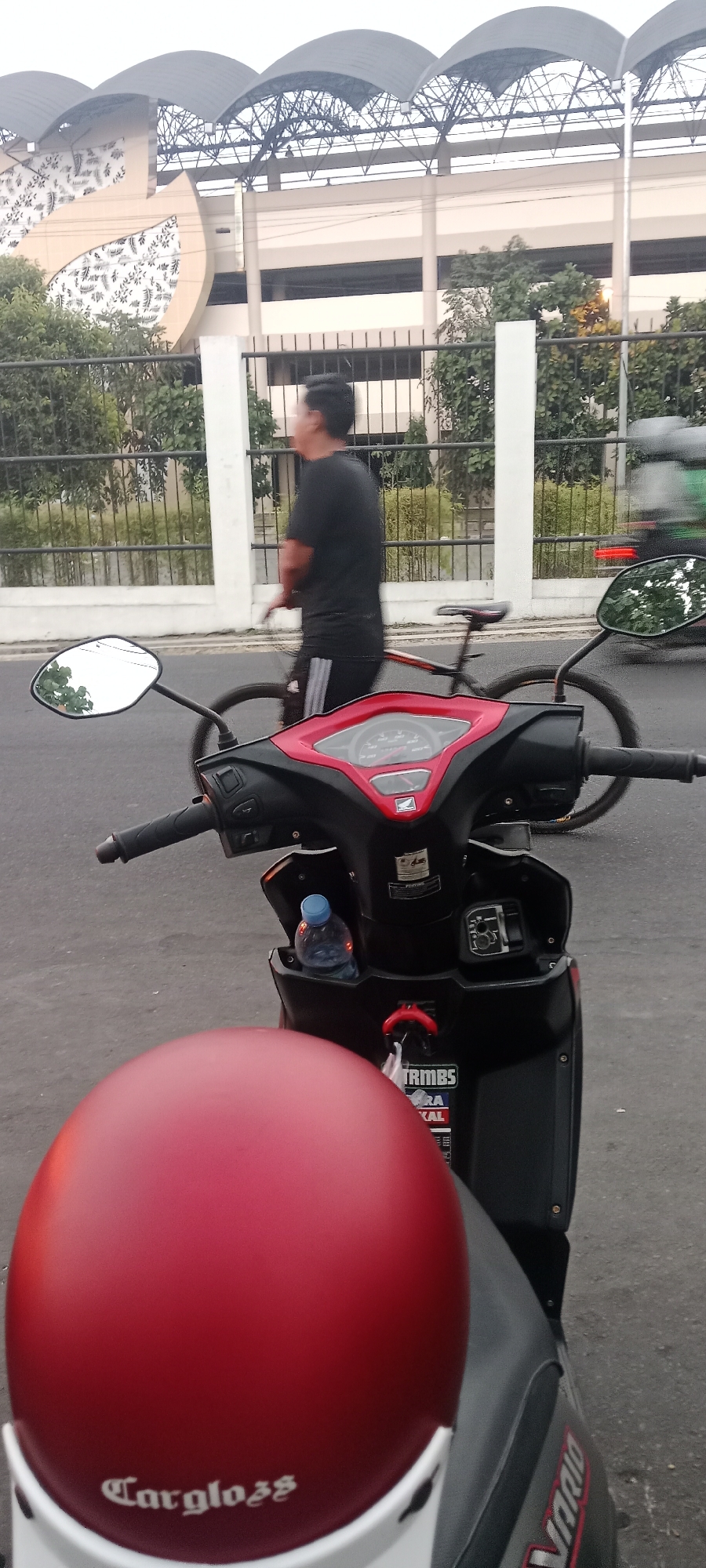 The image depicts a street scene in Ngemplak, Indonesia. In the foreground, a red motorcycle is parked, with its handlebars and instrument panel clearly visible. A red helmet rests on the seat of the motorcycle. In the midground, a man is walking alongside a bicycle on the sidewalk. Behind him, a fence separates the sidewalk from a building with a distinctive curved roof structure. The building appears to be a stadium or large public facility. In the background, trees and foliage are visible. The overall scene suggests a typical urban environment with pedestrian and vehicular activity.